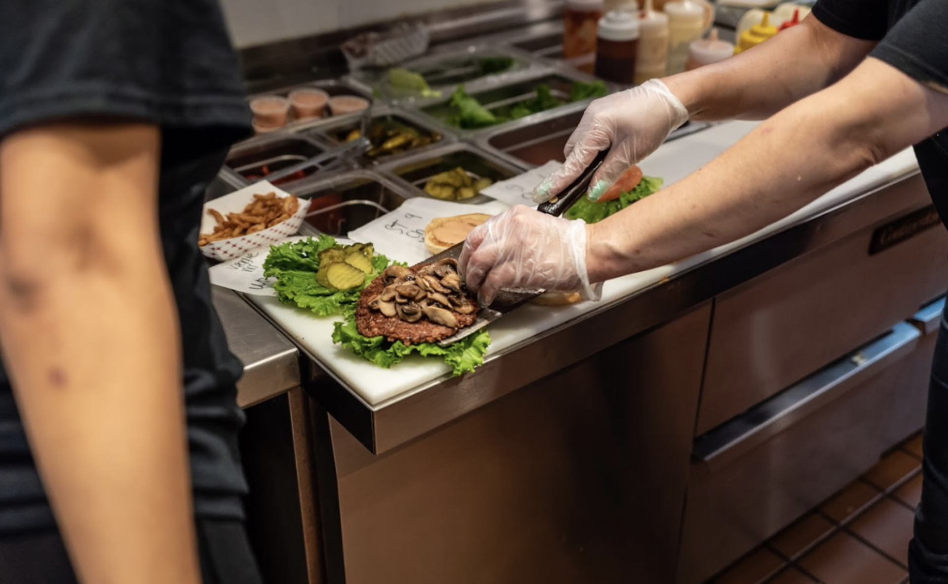 A person preparing a burger with lettuce, pickles, mushrooms, and other toppings at a restaurant kitchen.