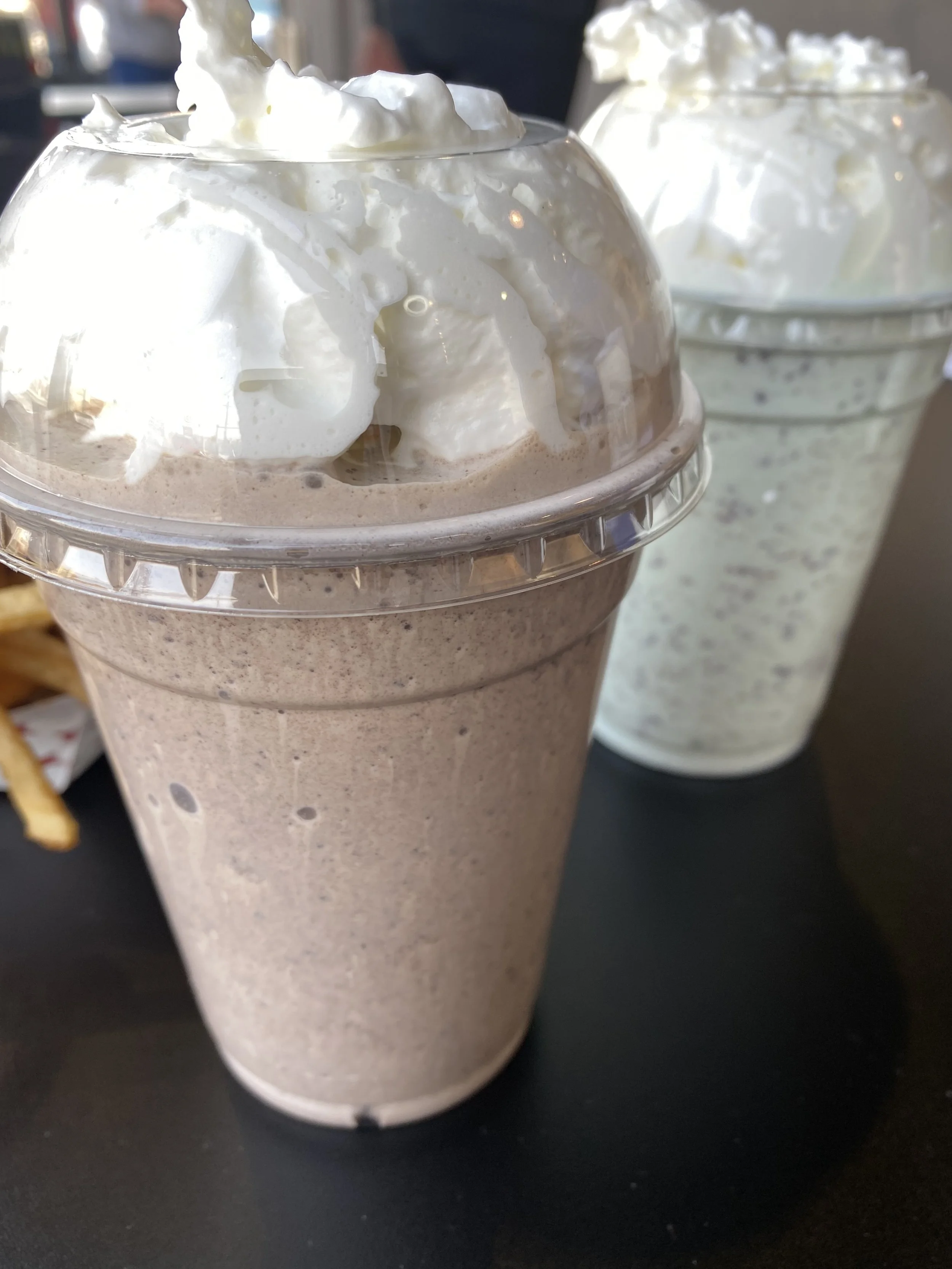 Two blended milkshakes topped with whipped cream, one chocolate and one cookies and cream, served in clear plastic cups with lids.