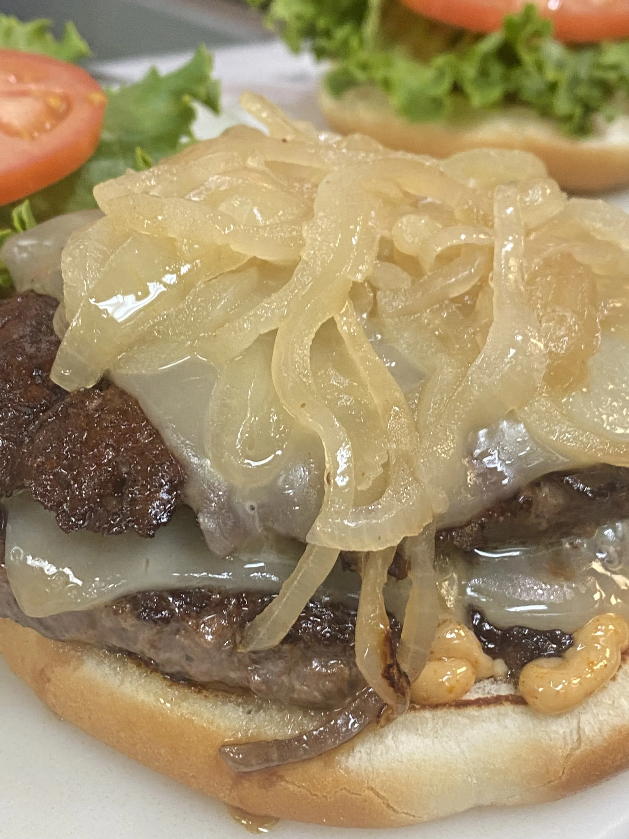 Close-up of a cheeseburger with caramelized onions, melted cheese, and a beef patty inside a bun, with tomato slices and lettuce in the background.