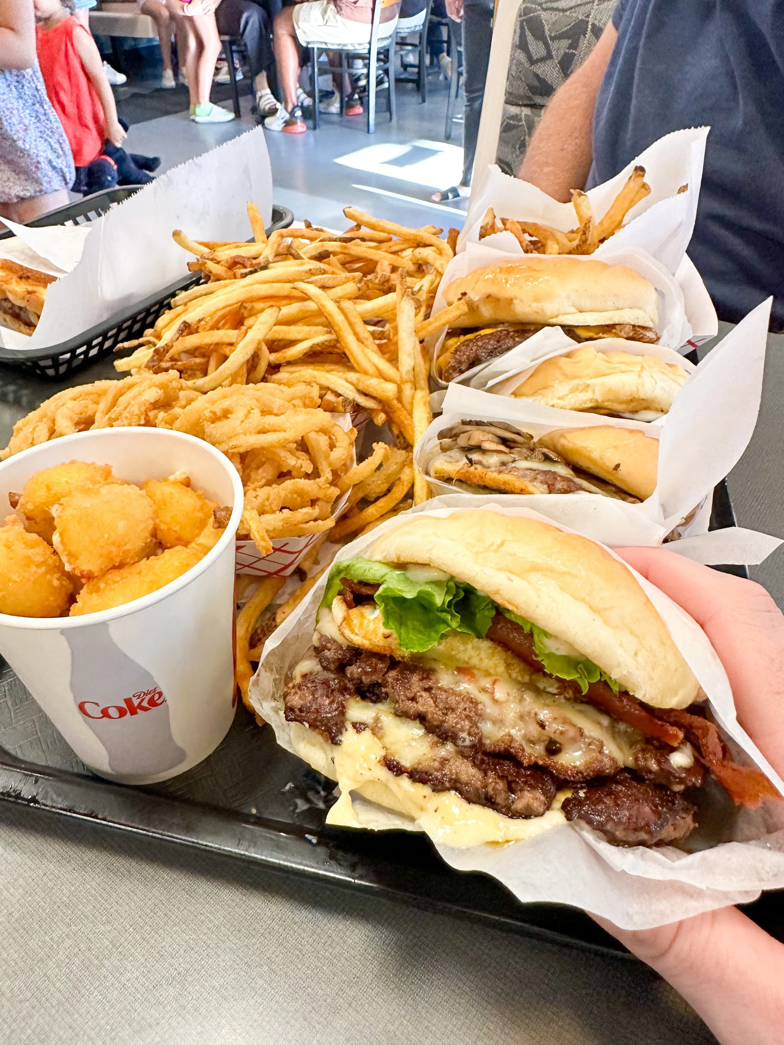 A tray of fast food including a cheeseburger with lettuce, tomato, and bacon, several other burgers, French fries, onion rings, and a cup of fried chicken bites inside a casual restaurant.