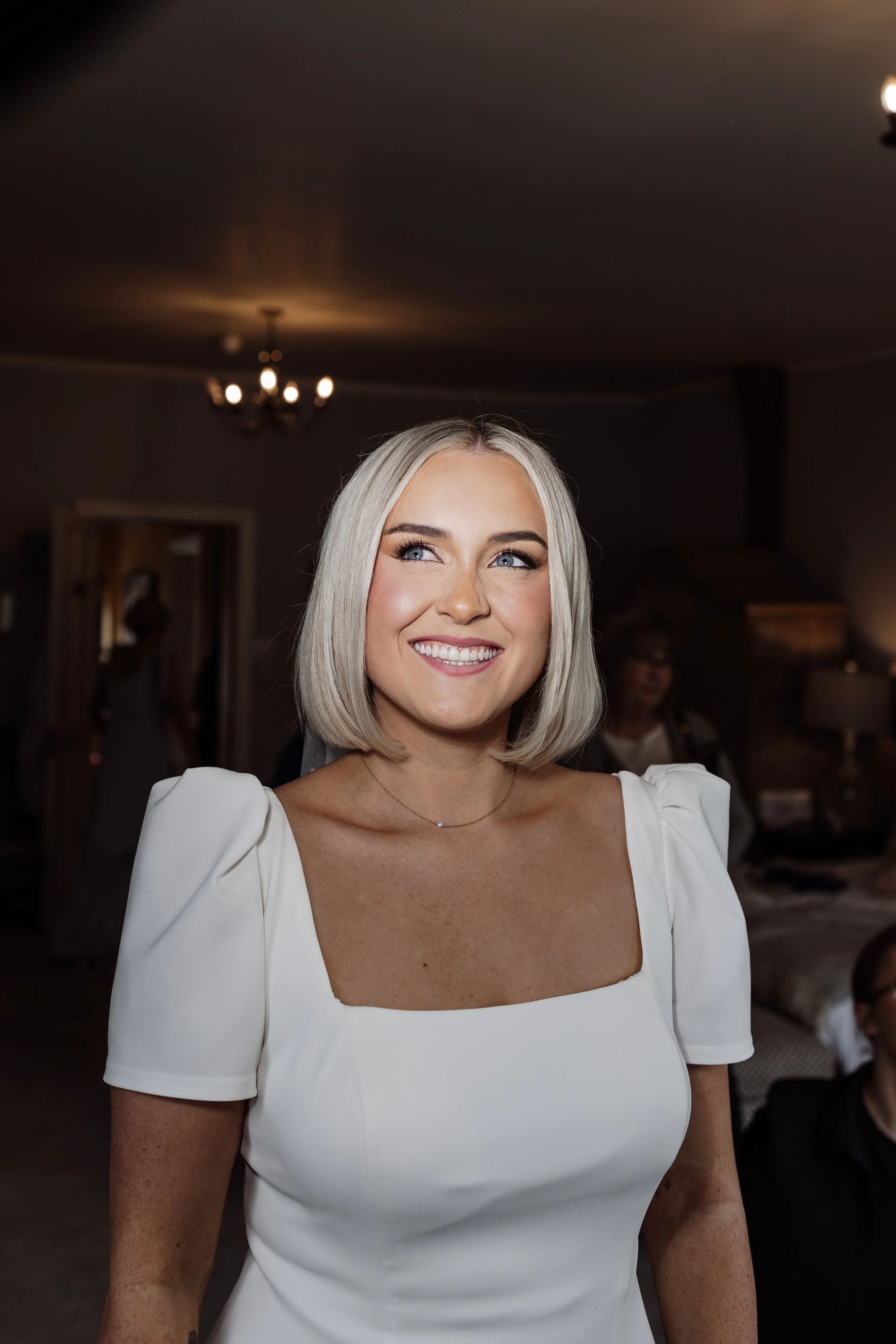 Happy woman with blonde bob haircut wearing a white wedding dress with puff sleeves and a delicate necklace, indoors with dim lighting and blurry background.