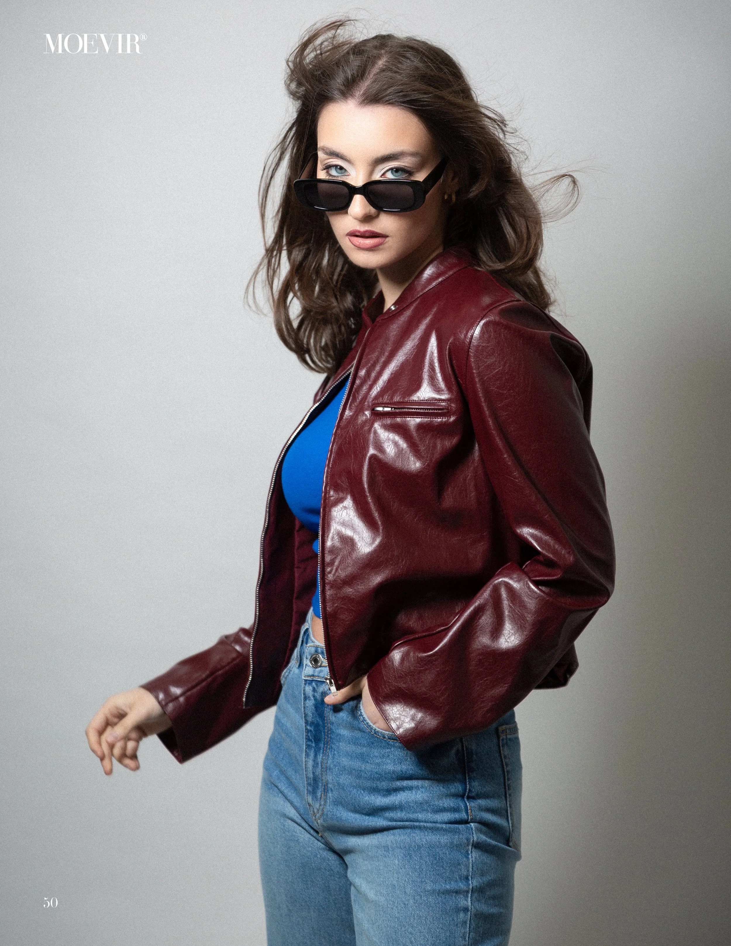 A young woman with wavy brown hair wearing black sunglasses, a maroon leather jacket, a blue top, and light blue jeans, standing against a plain light gray background.
