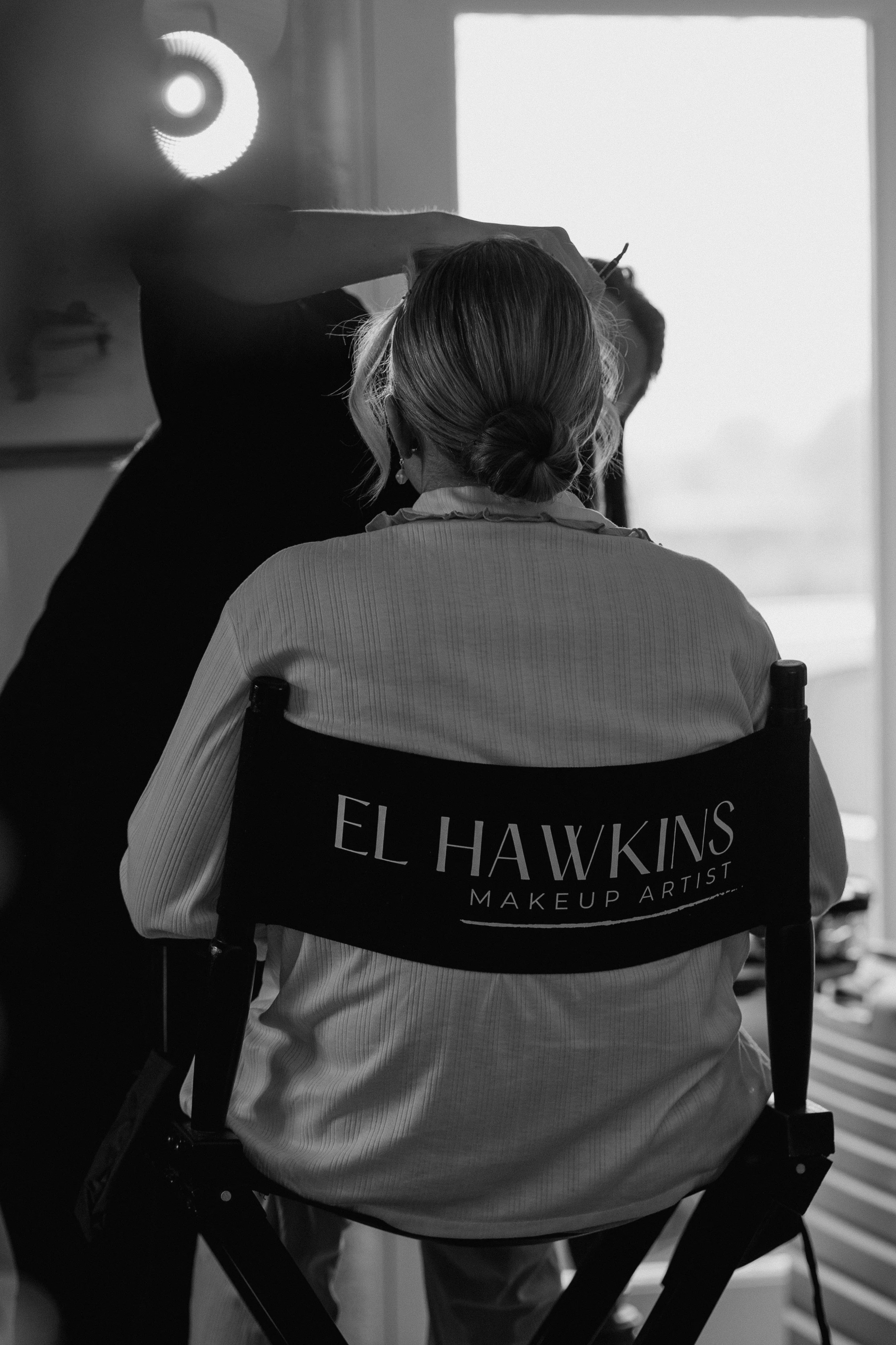 A person sitting in a makeup chair with 'El Hawkins Makeup Artist' printed on the back, getting makeup applied by a stylist, with natural light coming through a window.
