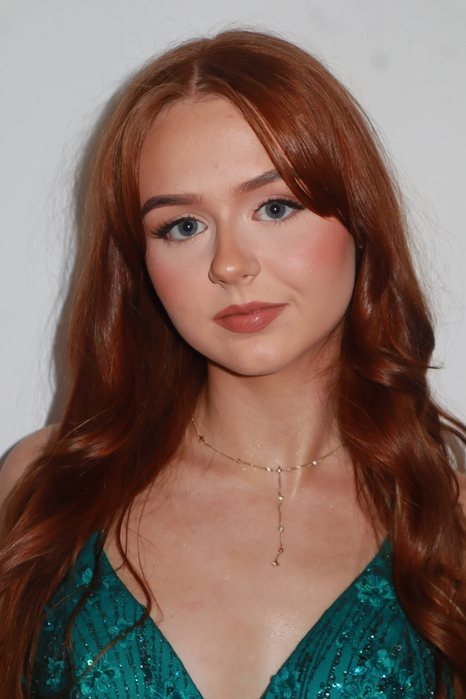 A young woman with long red hair, blue eyes, and fair skin. She is wearing makeup, a teal dress with floral patterns, and a delicate gold necklace. She is looking directly at the camera with a slight smile.