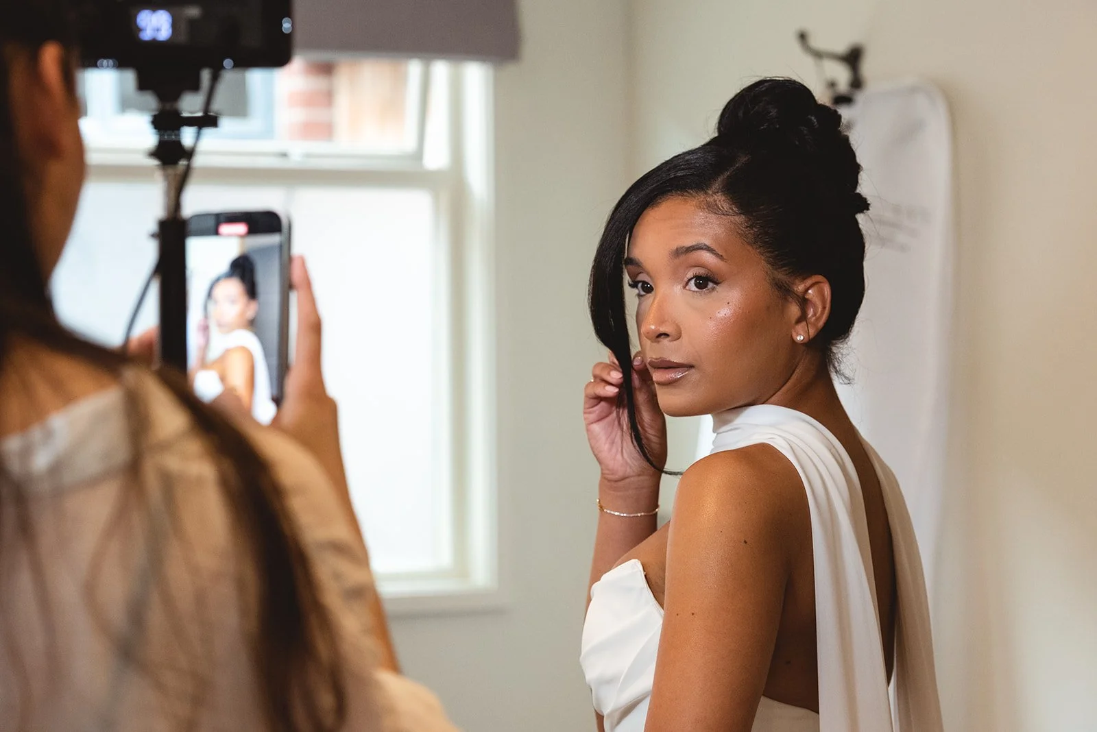 A woman with dark hair styled in an updo and wearing a white wedding dress, posing for a photograph indoors while another person takes her picture with a smartphone.