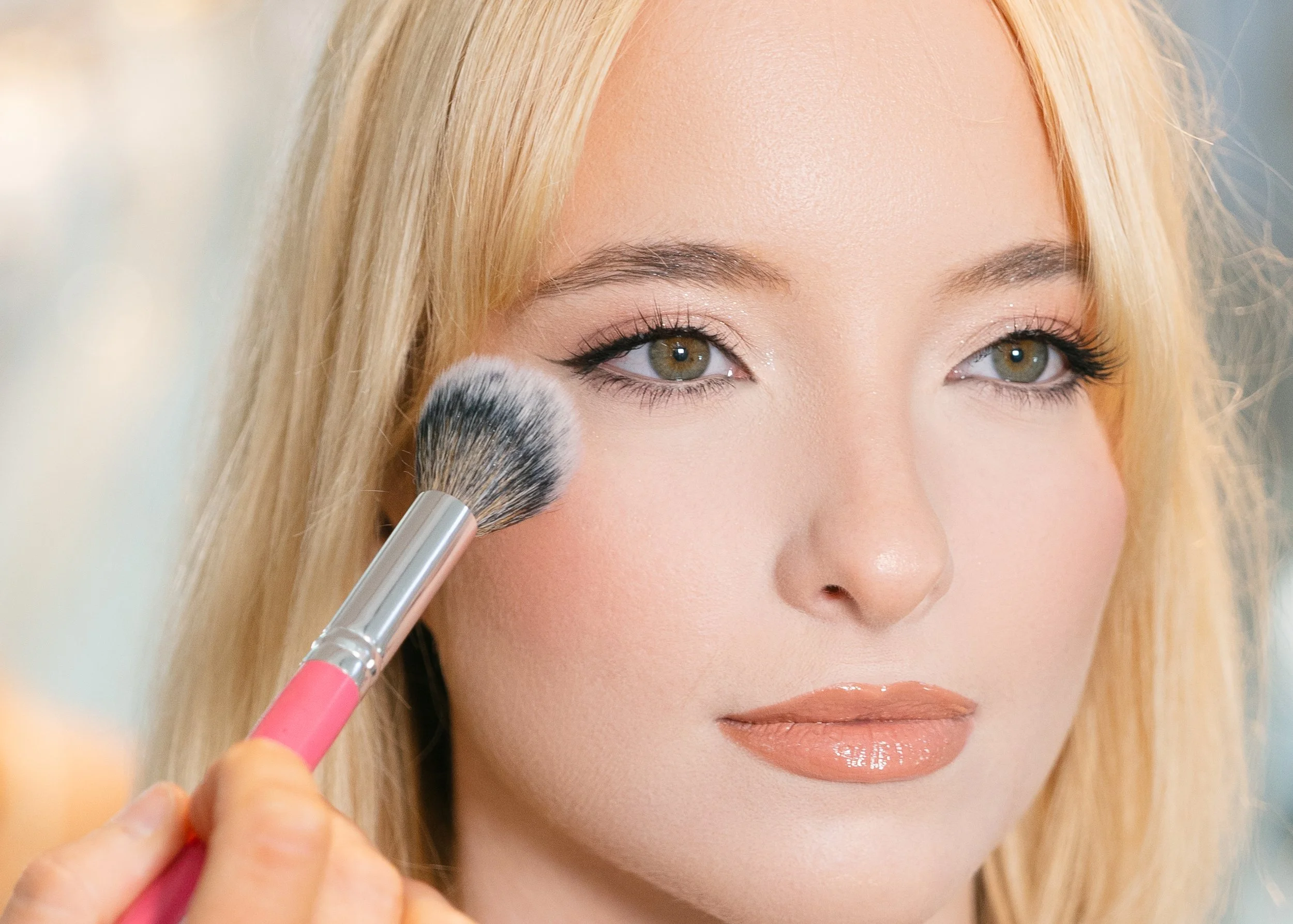 A woman with blonde hair and makeup is applying blush or powder to her cheek with a makeup brush.