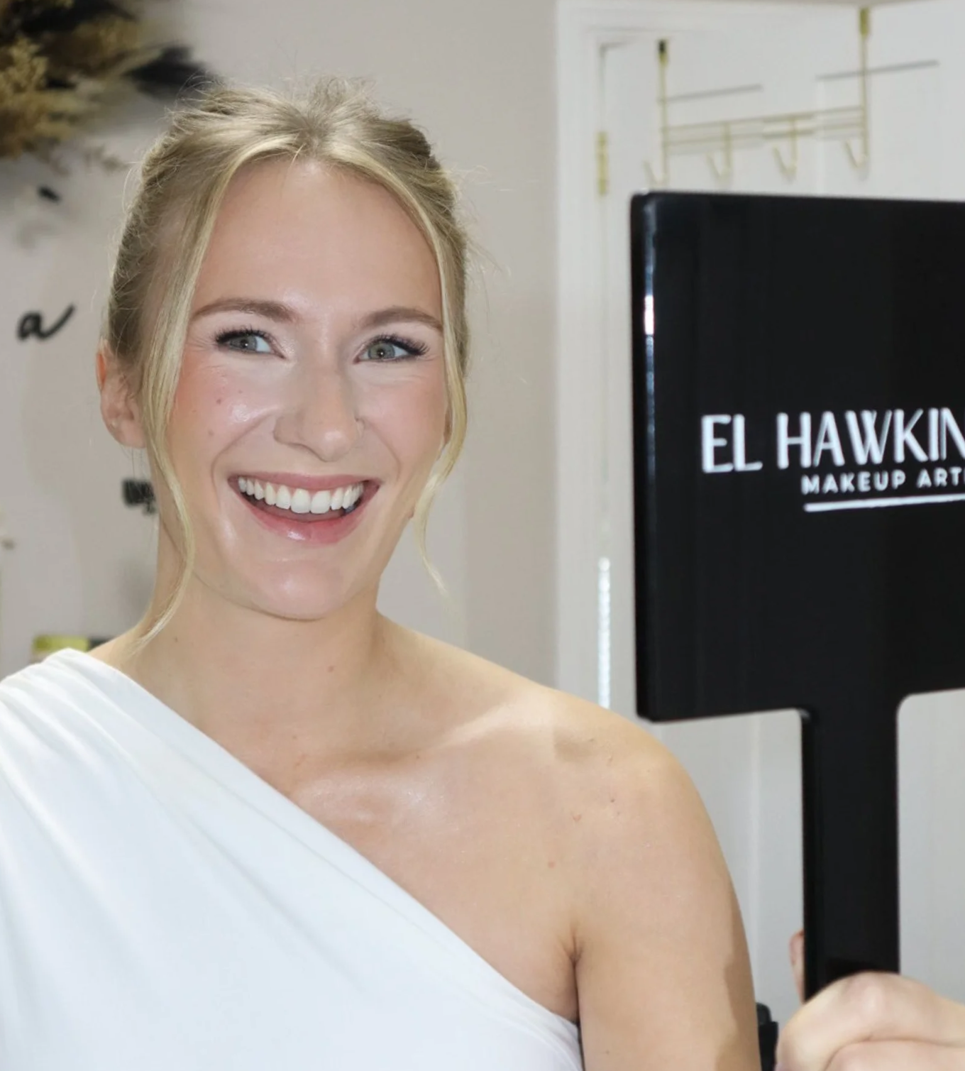 A smiling woman holding a mirror with 'El Hawkin Makeup Art' written on it.