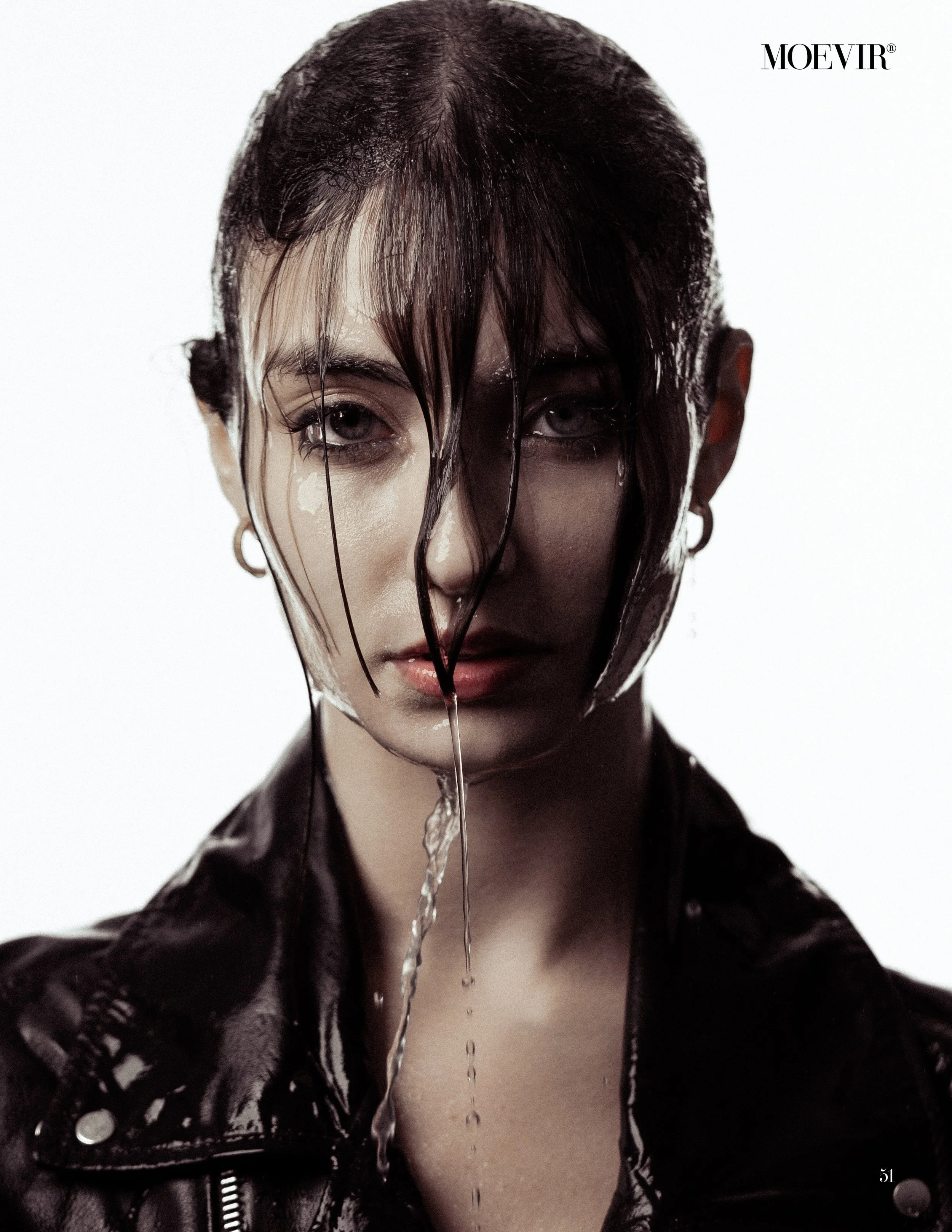 Portrait of a woman with wet hair and water dripping from her face, wearing a black leather jacket, against a plain white background.