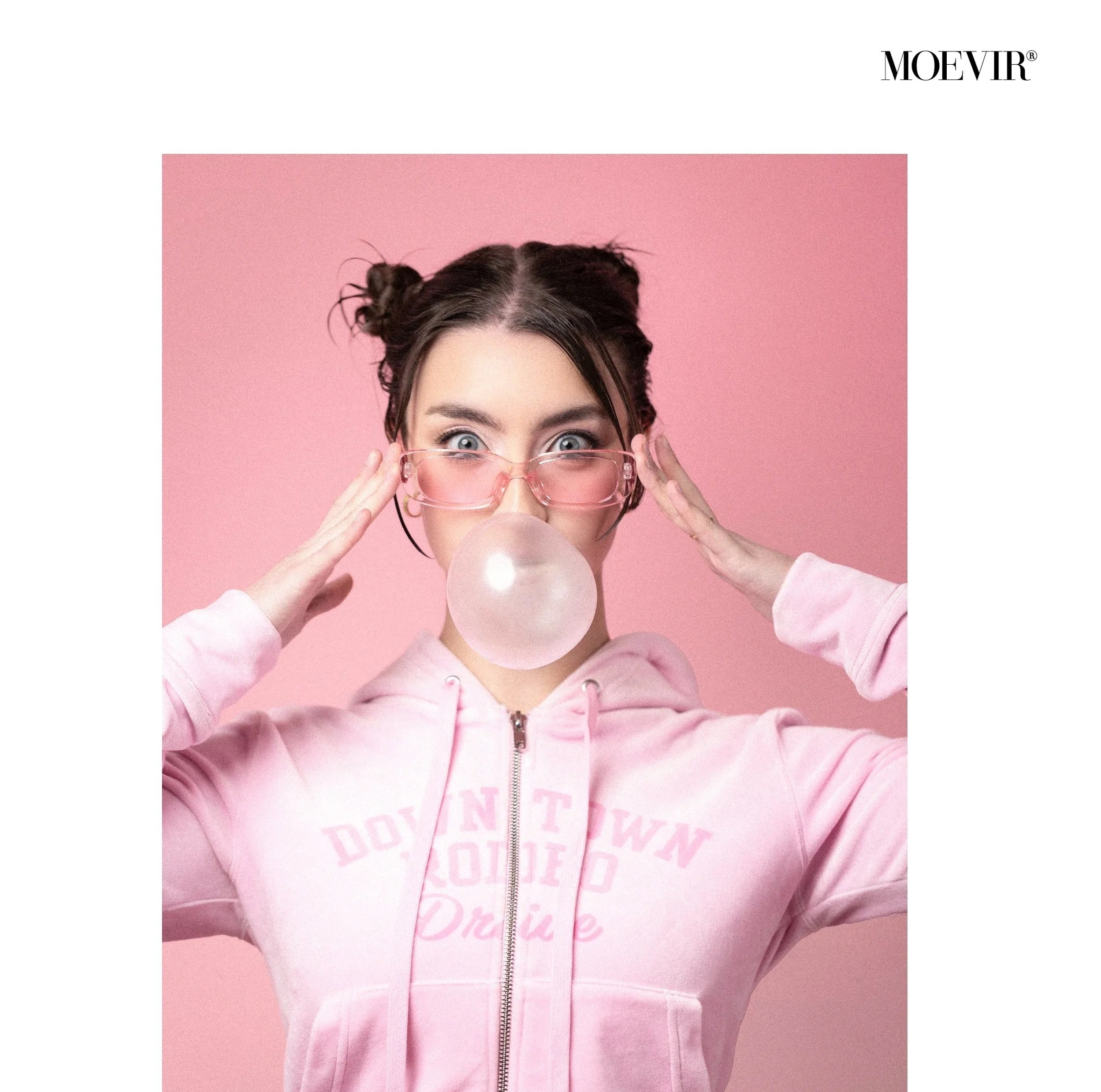 A young woman with dark hair styled in two buns, pink-tinted glasses, and a pink hoodie blows a bubble with gum against a pink background.