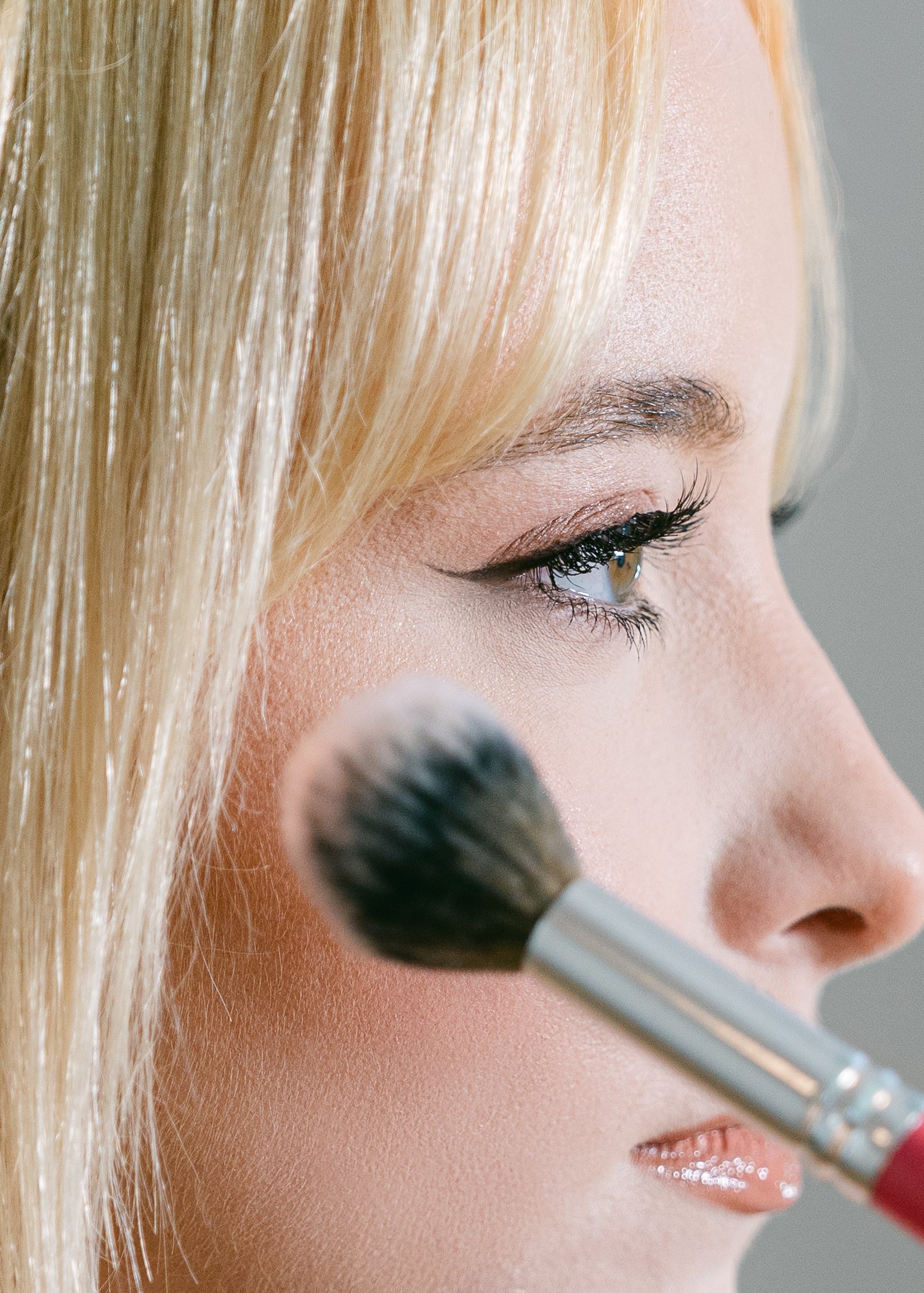 Close-up of a woman with blonde hair applying makeup using a fluffy brush, with her eye makeup visible and soft lighting.