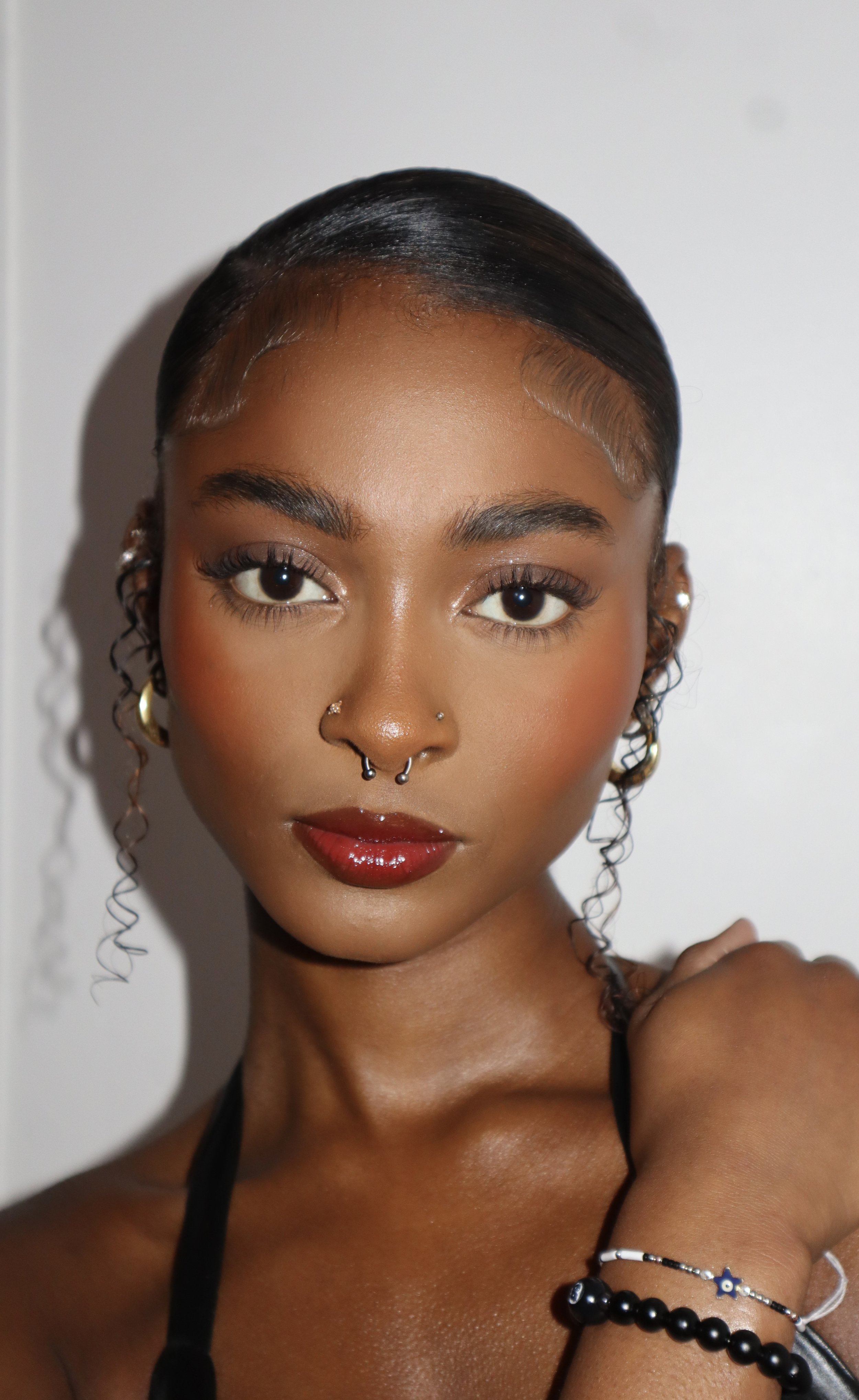 A young woman with dark, sleek, pulled-back hair, gold jewelry, and bright makeup looking at the camera.