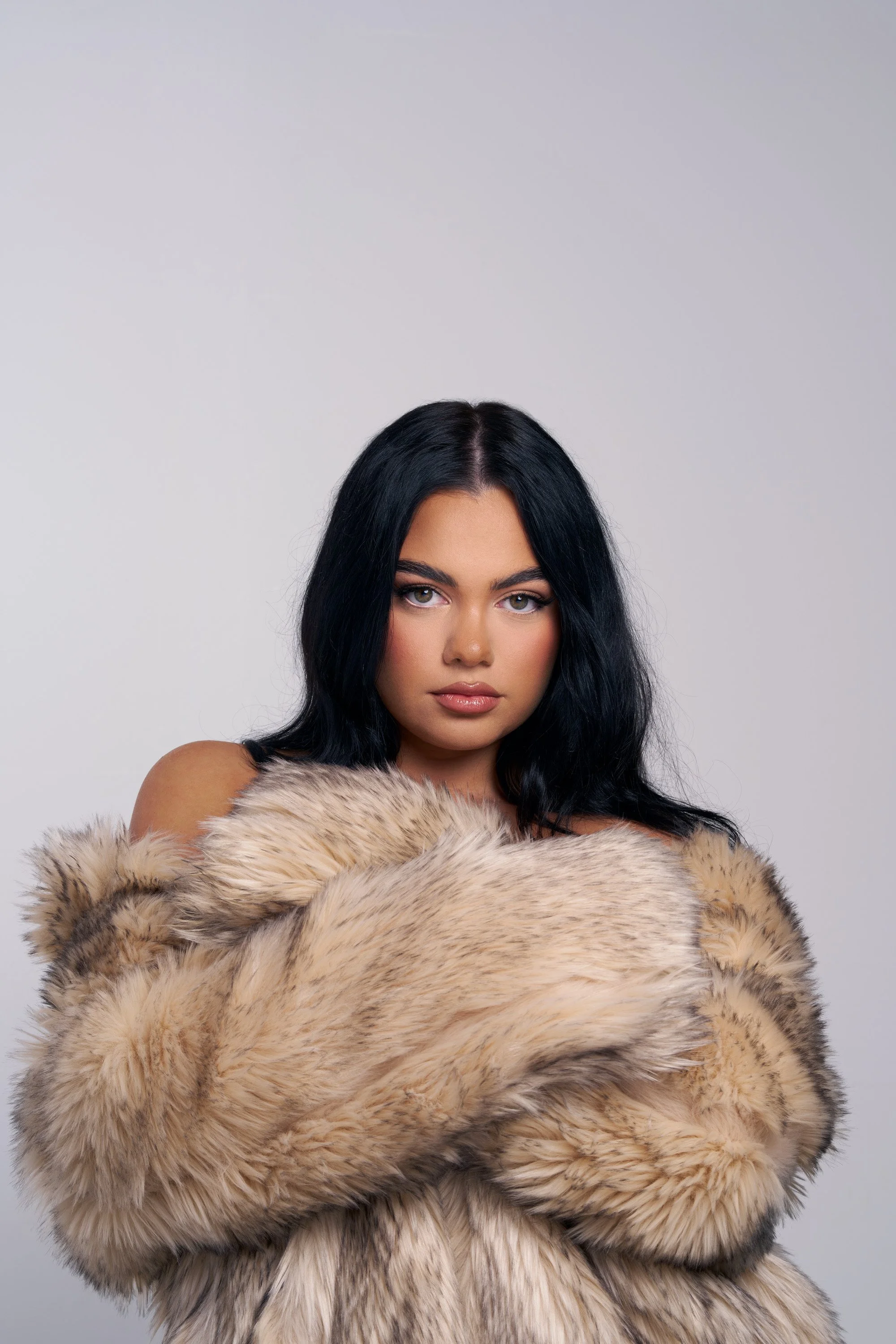 A woman with long black hair, light skin, and makeup, wearing a large beige and brown fur coat, standing against a plain gray background.
