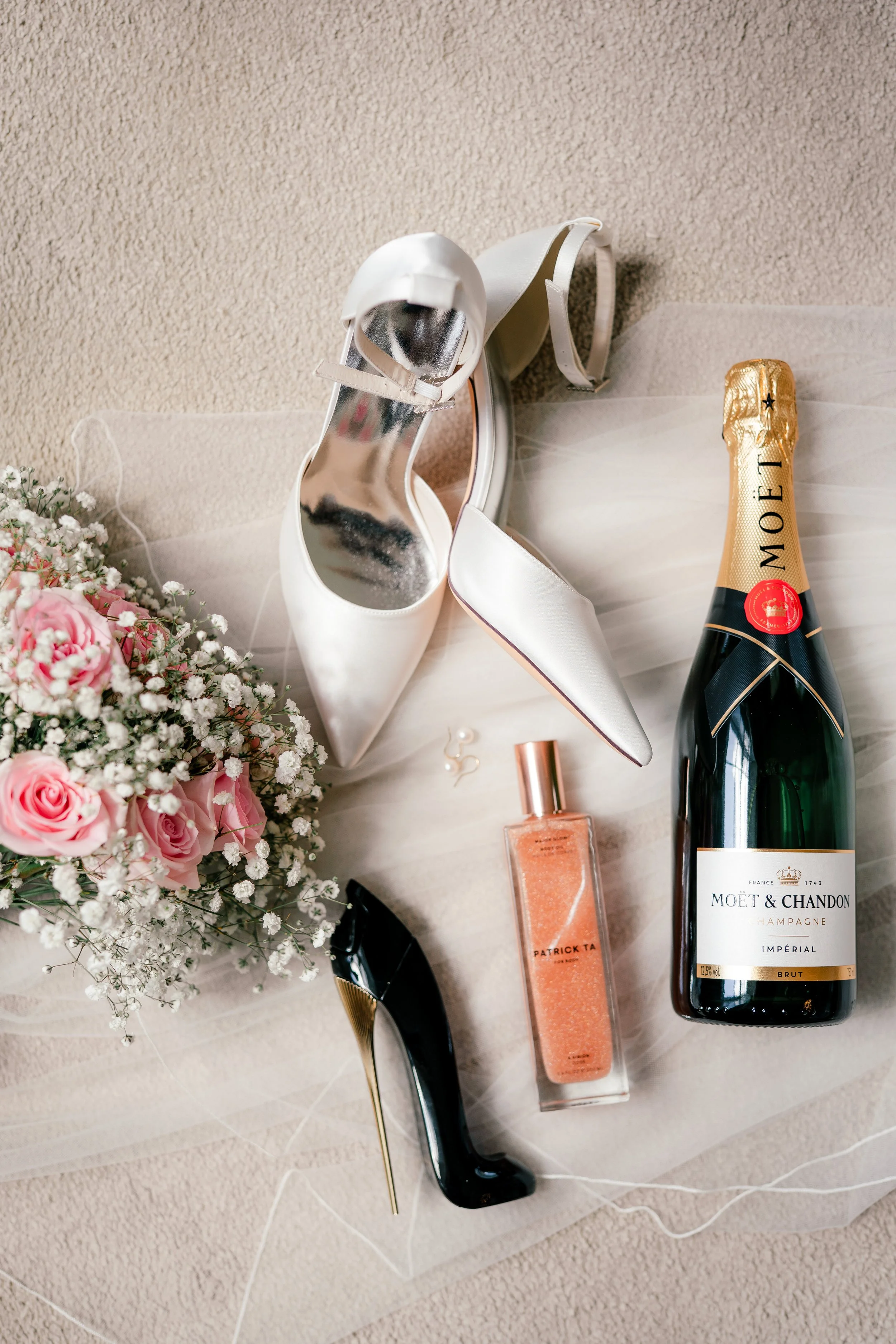 Elegant white high heels, black high heels, a bouquet of pink and white flowers, a bottle of Moët & Chandon champagne, a bottle of peach-colored perfume, and pearl earrings arranged on a light-colored surface.