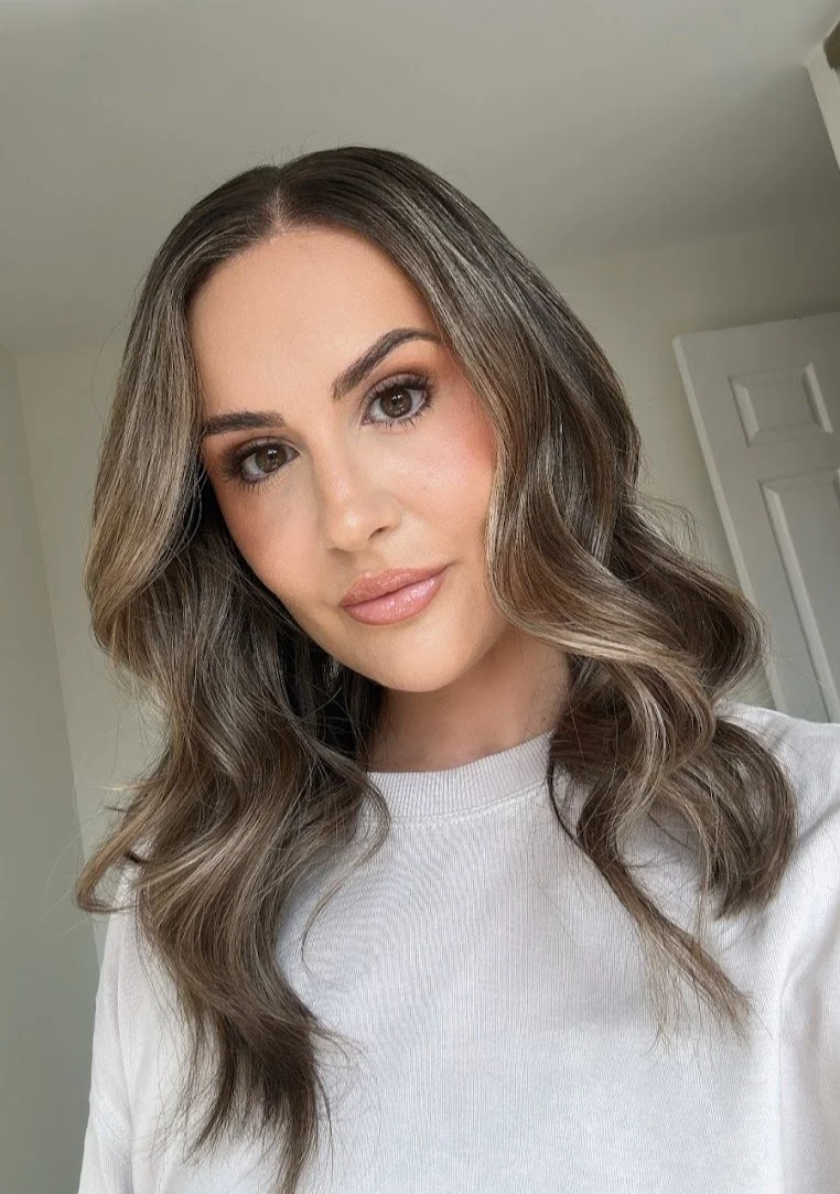A woman with wavy, shoulder-length brown hair and brown eyes, wearing a white shirt, standing indoors with a door in the background.