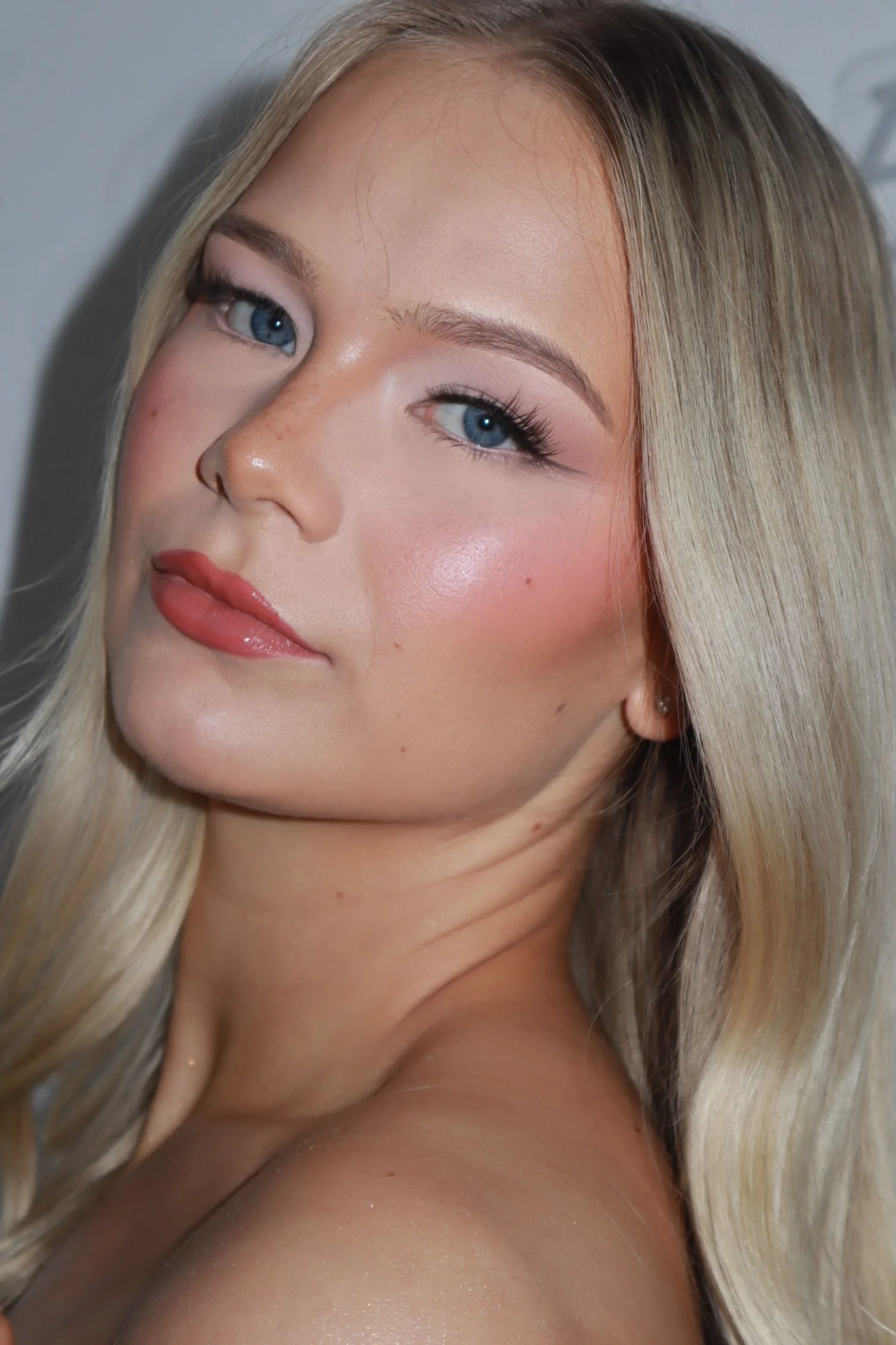 Close-up of a young woman with long blonde hair and blue eyes, wearing makeup with pink blush and lipstick, softly smiling indoors.
