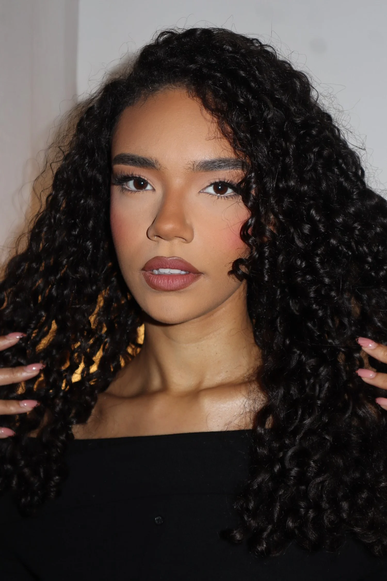Close-up of a young woman with curly black hair, brown eyes, and wearing a black top, looking directly at the camera.
