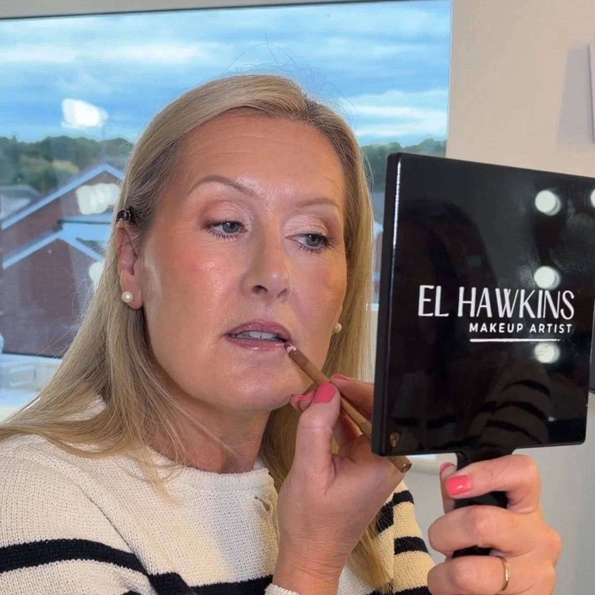 A woman with blonde hair and pearl earrings applying makeup with a pencil, looking into a mirror labeled 'El Hawkins Makeup Artist'.