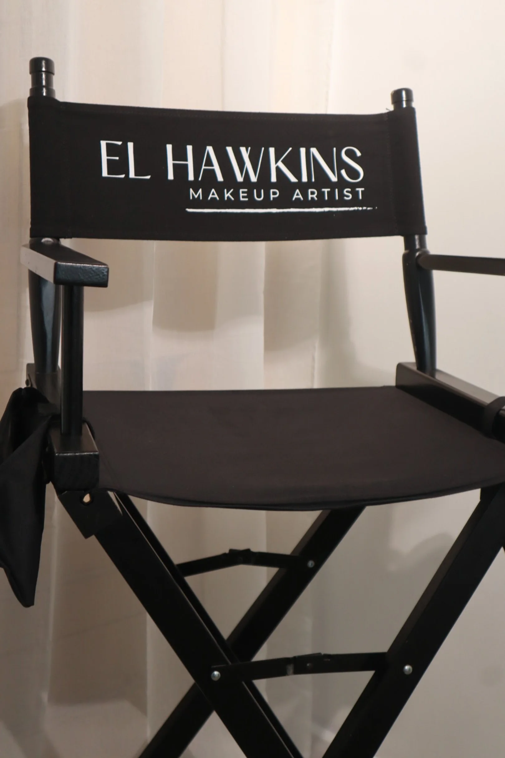 Black director's chair with white text that reads 'El Hawkins Makeup Artist' against a cream-colored curtain background.