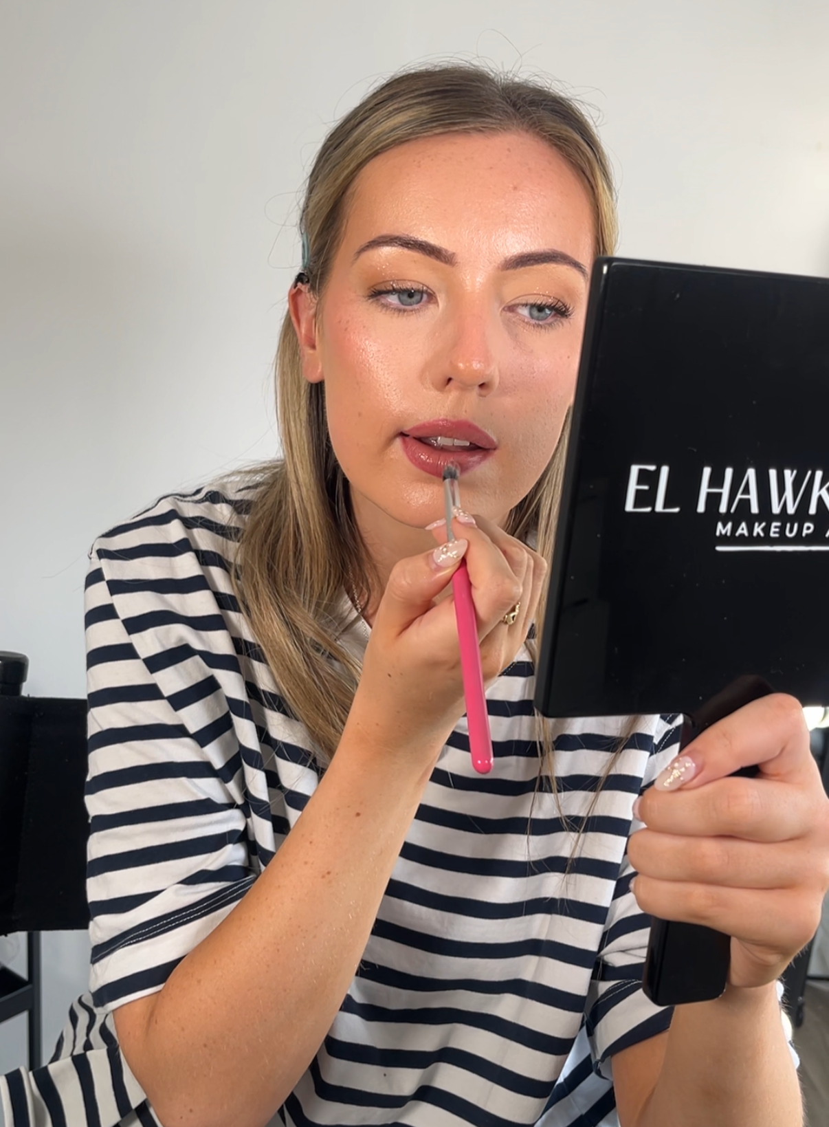 Woman applying lipstick while looking into a mirror labeled 'El Hawke Makeup'.