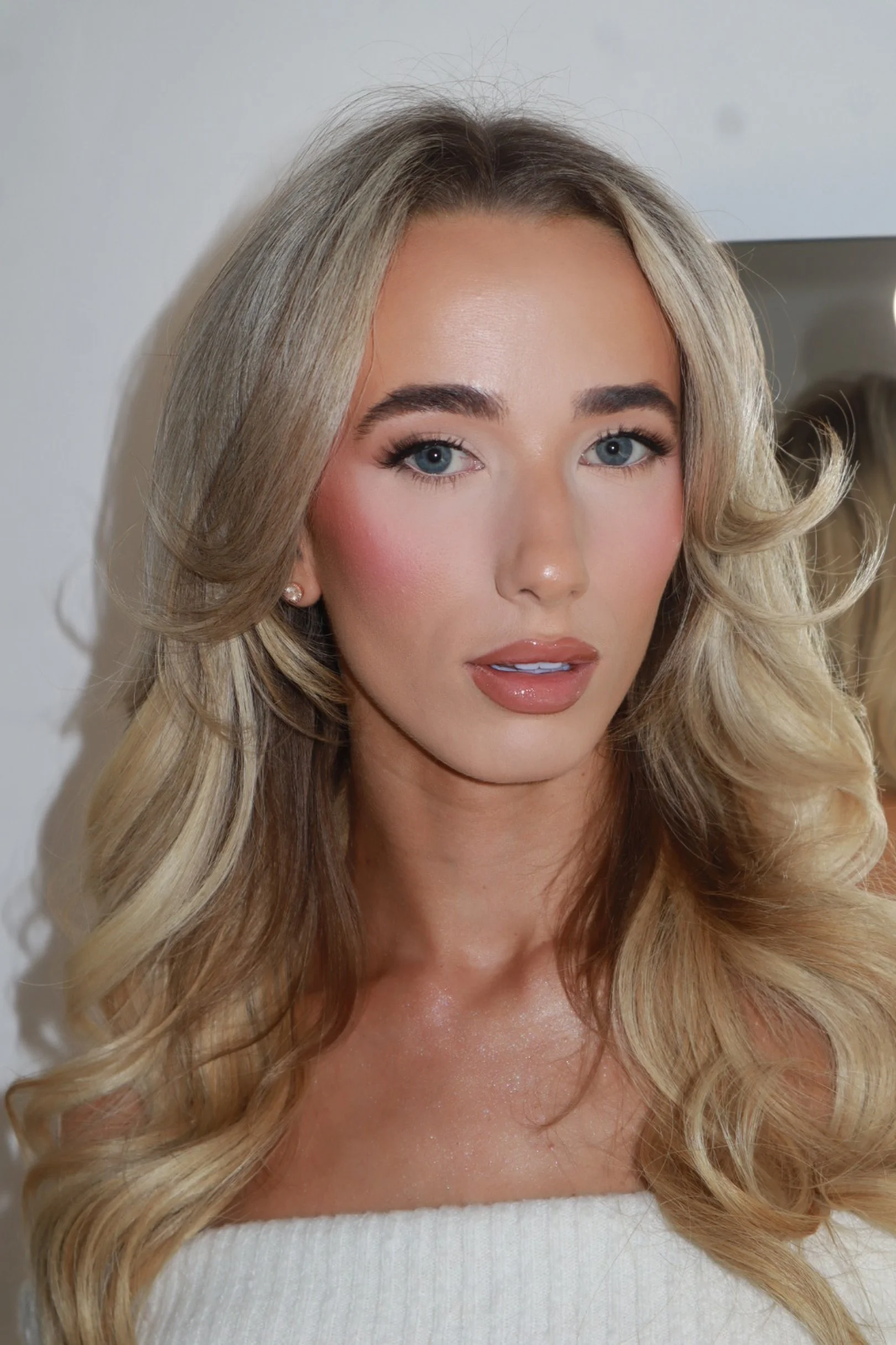 Close-up of a woman with long wavy blonde hair, blue eyes, and makeup, wearing a white top and pearl earrings, standing in front of a mirror.