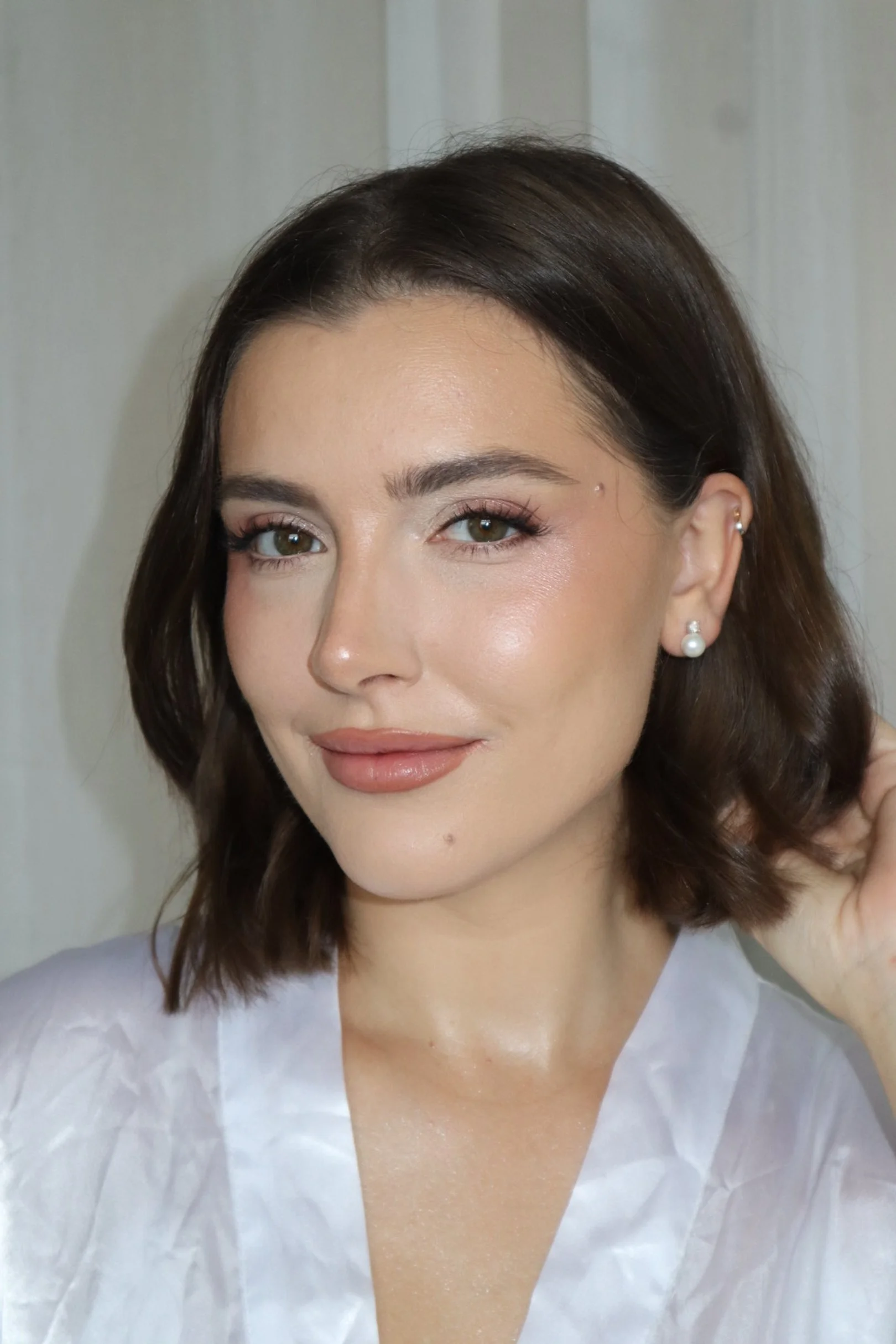 A woman with brown hair, green eyes, makeup, and pearl earrings posing indoors.
