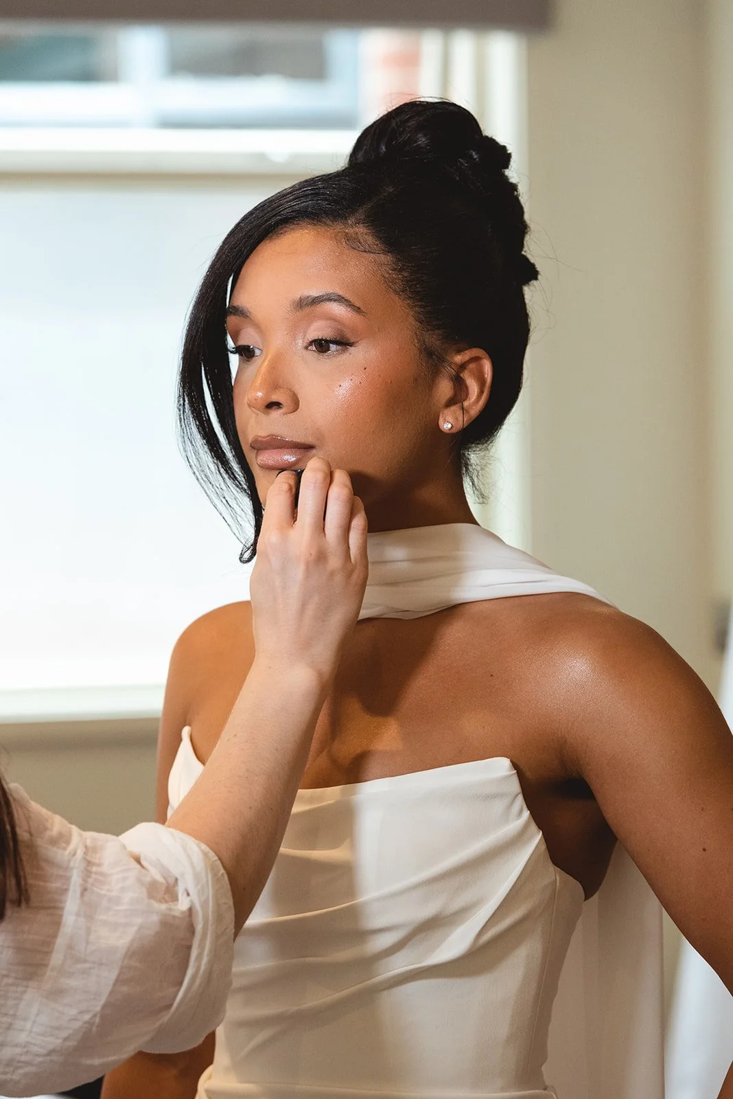 A bride with dark hair styled in an updo, wearing a strapless white dress, is having makeup applied by a makeup artist, with natural light coming from a window in the background.
