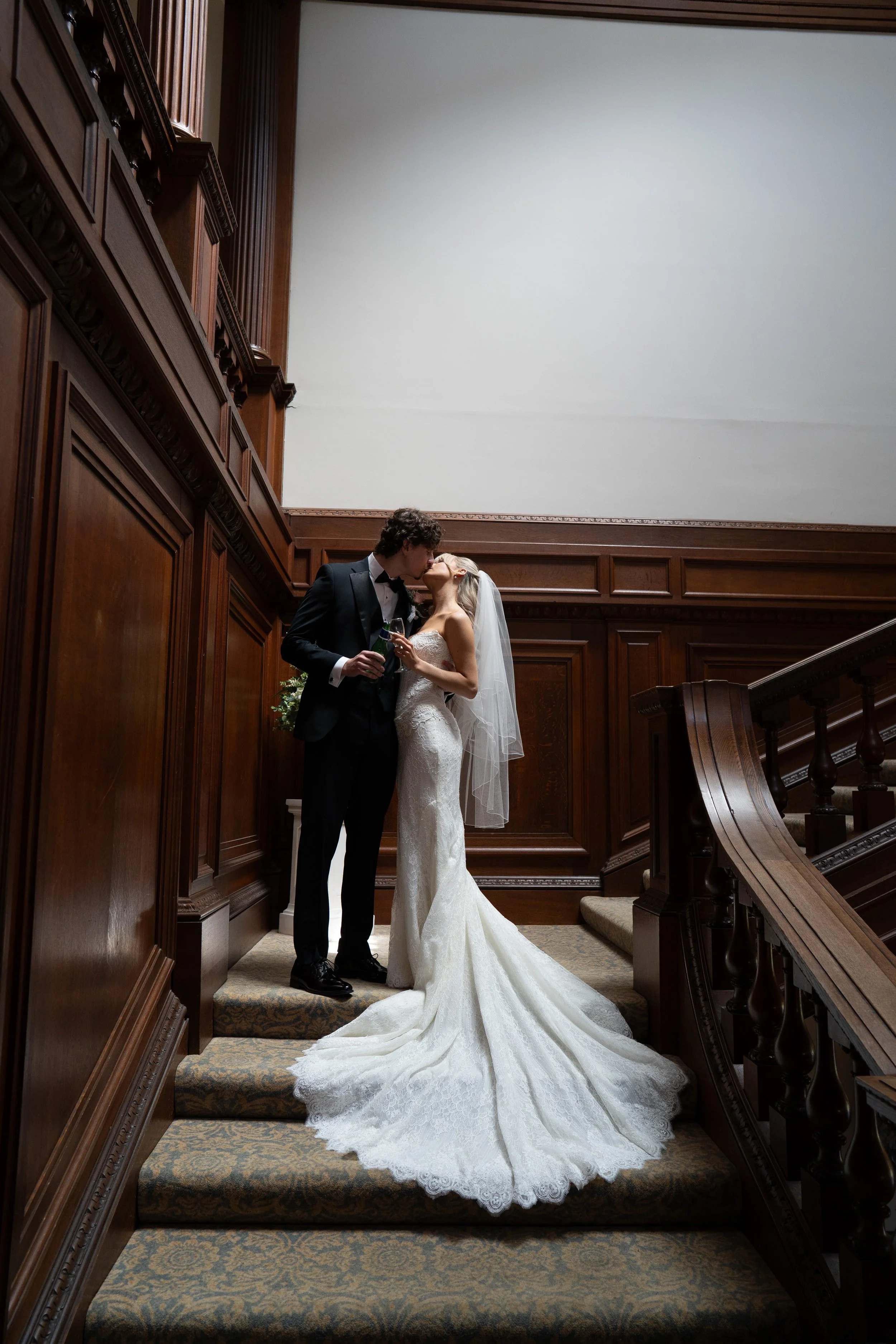 A bride and groom sharing a kiss on a staircase in a wood-paneled interior, with the bride in a white wedding gown and veil, and the groom in a black tuxedo, holding a glass of champagne.