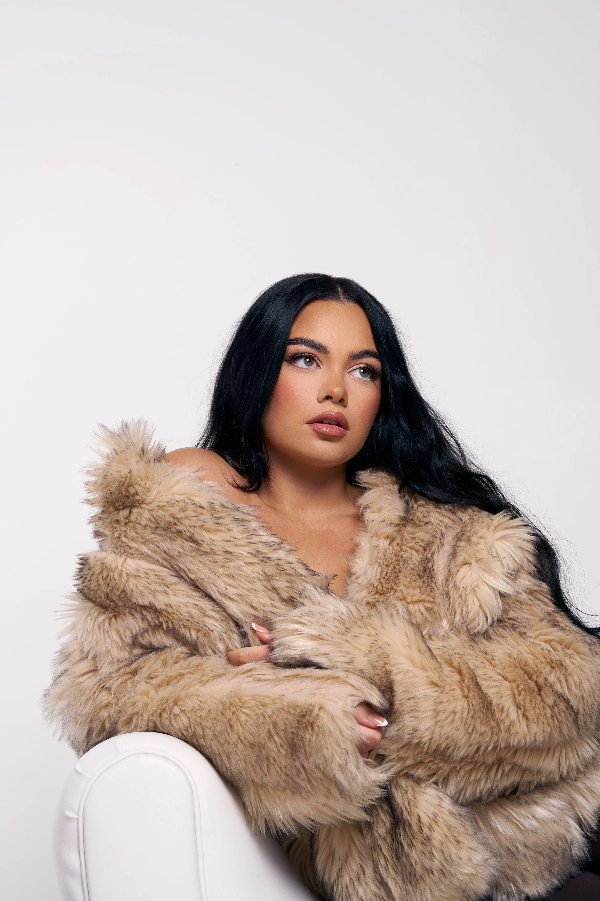 A woman with long black hair and makeup, wearing a large beige fur coat, sitting on a white chair against a plain white background.