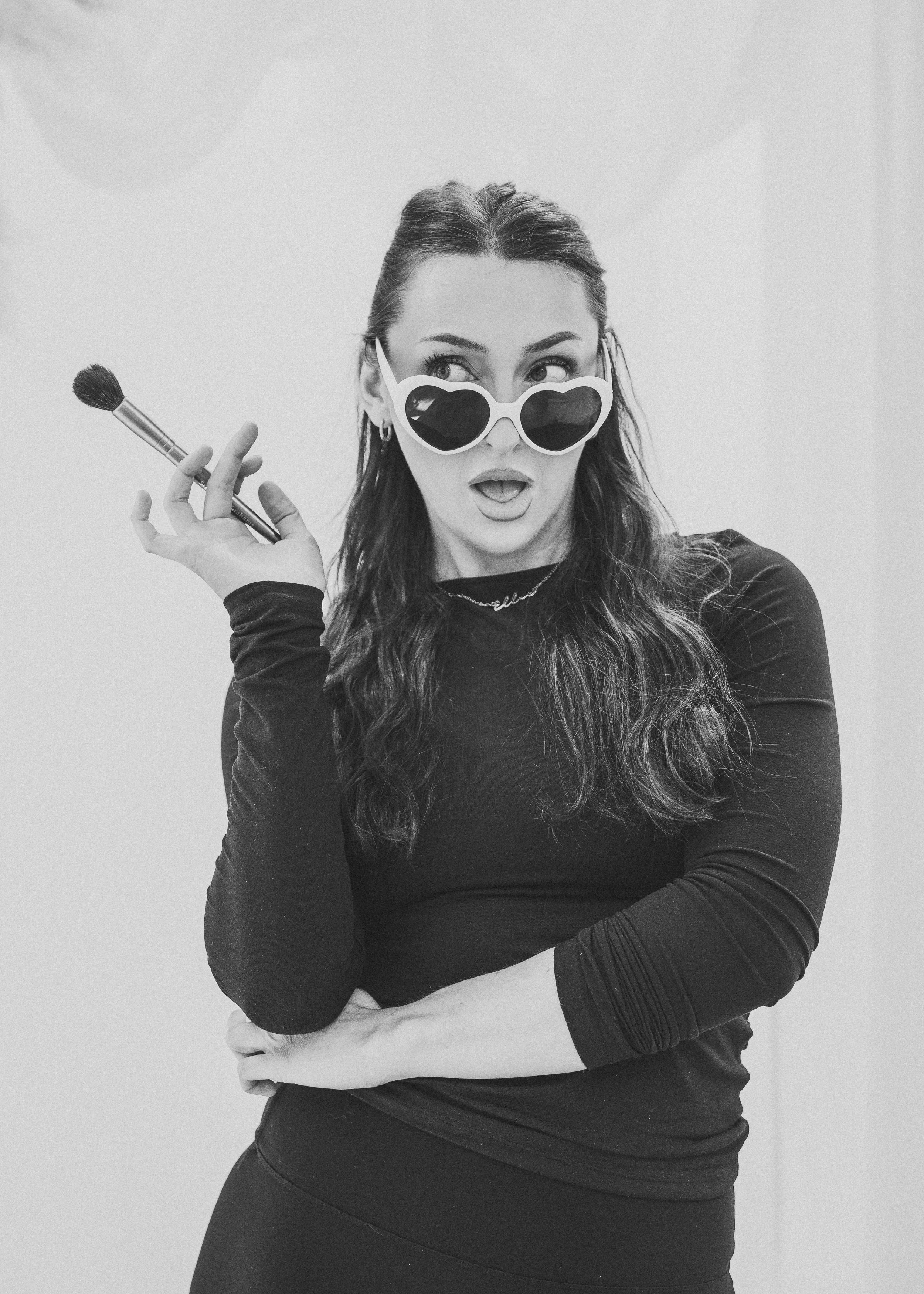 Bridal makeup artist with heart shaped glasses on with a shocked face holding a makeup brush