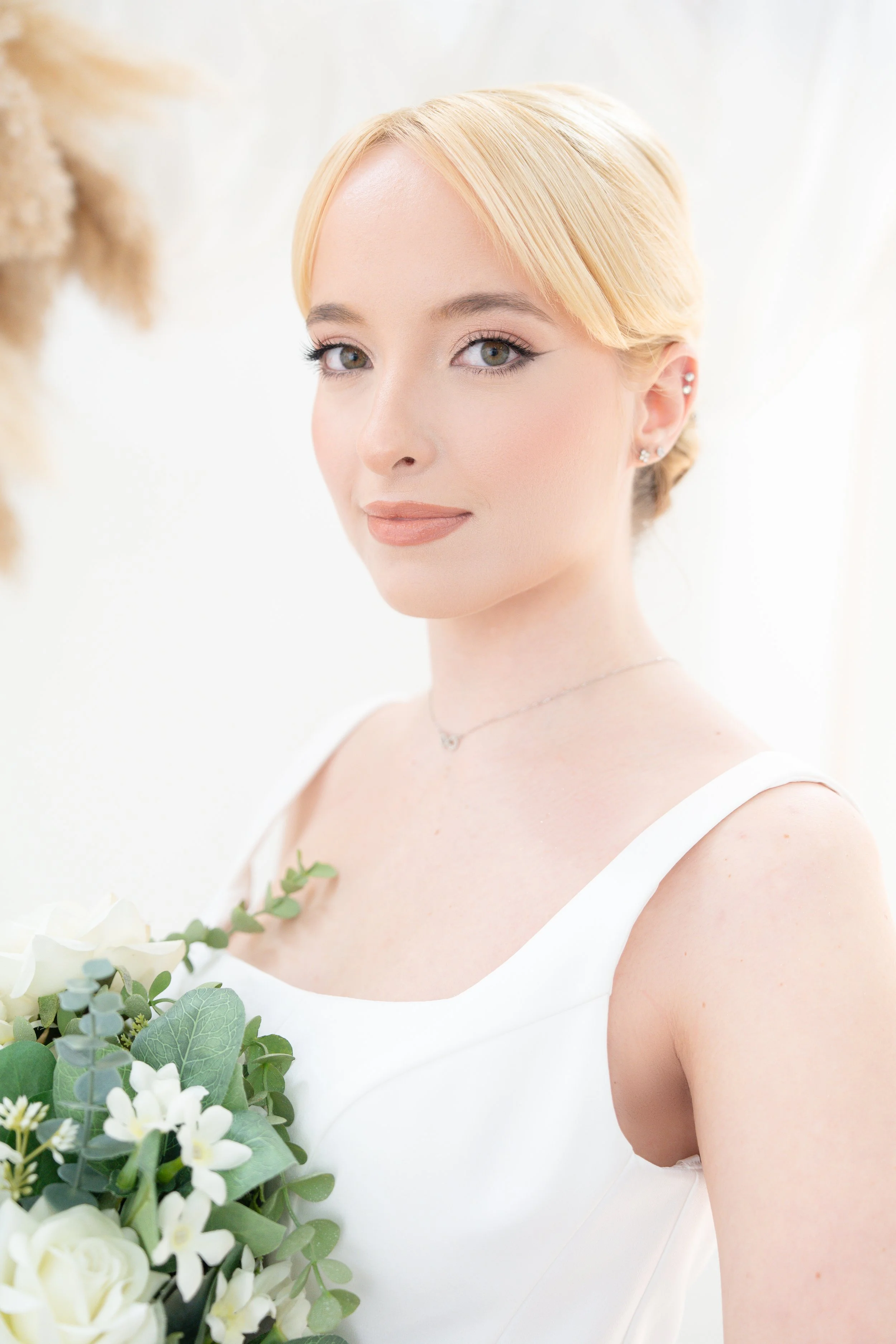 A young woman in a white dress holding a bouquet of white and green flowers, with blonde hair styled in an elegant updo, standing indoors with soft lighting.