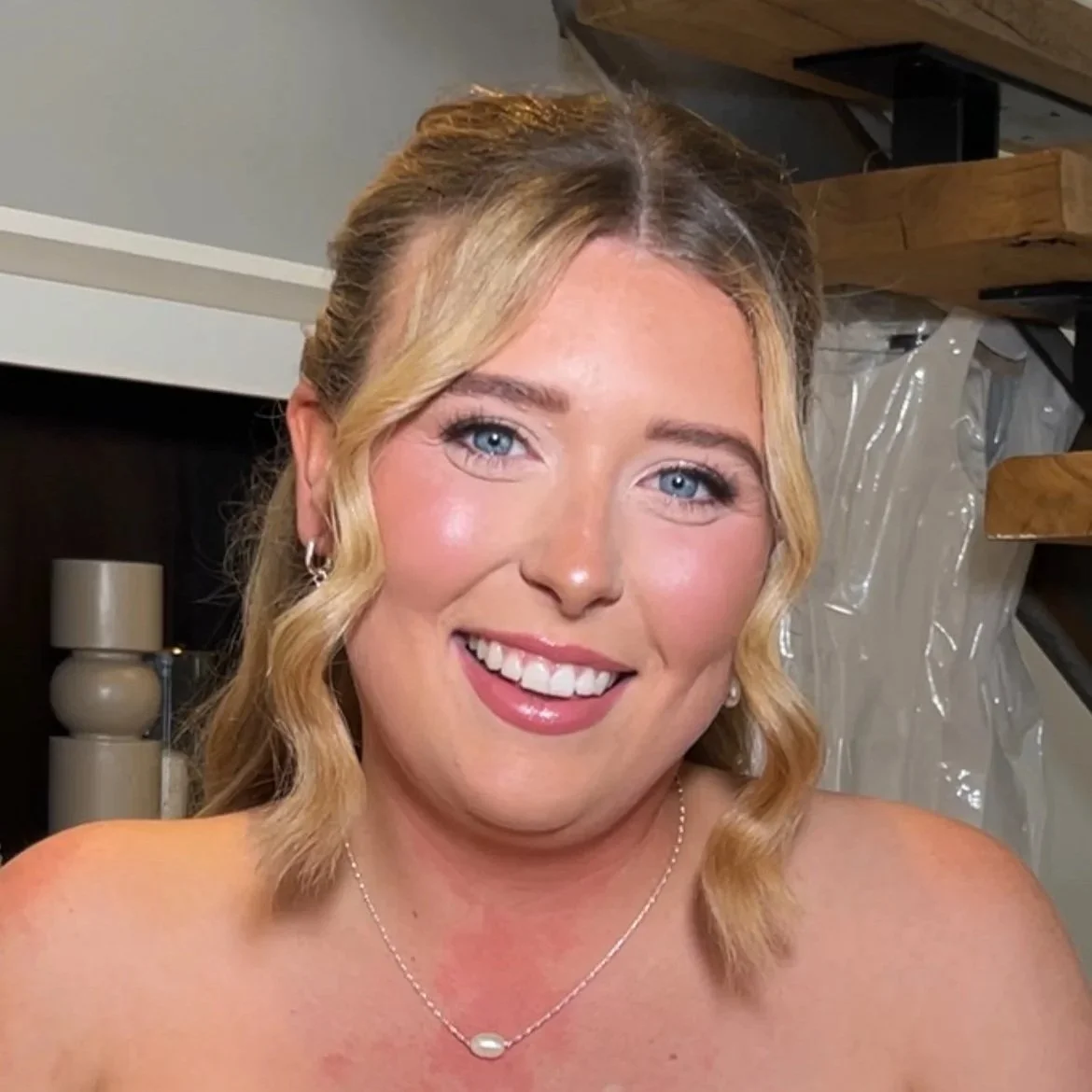 A smiling woman with blonde hair styled in loose waves, wearing a pearl necklace and earrings, indoors against a background of wooden stairs and a white backdrop.