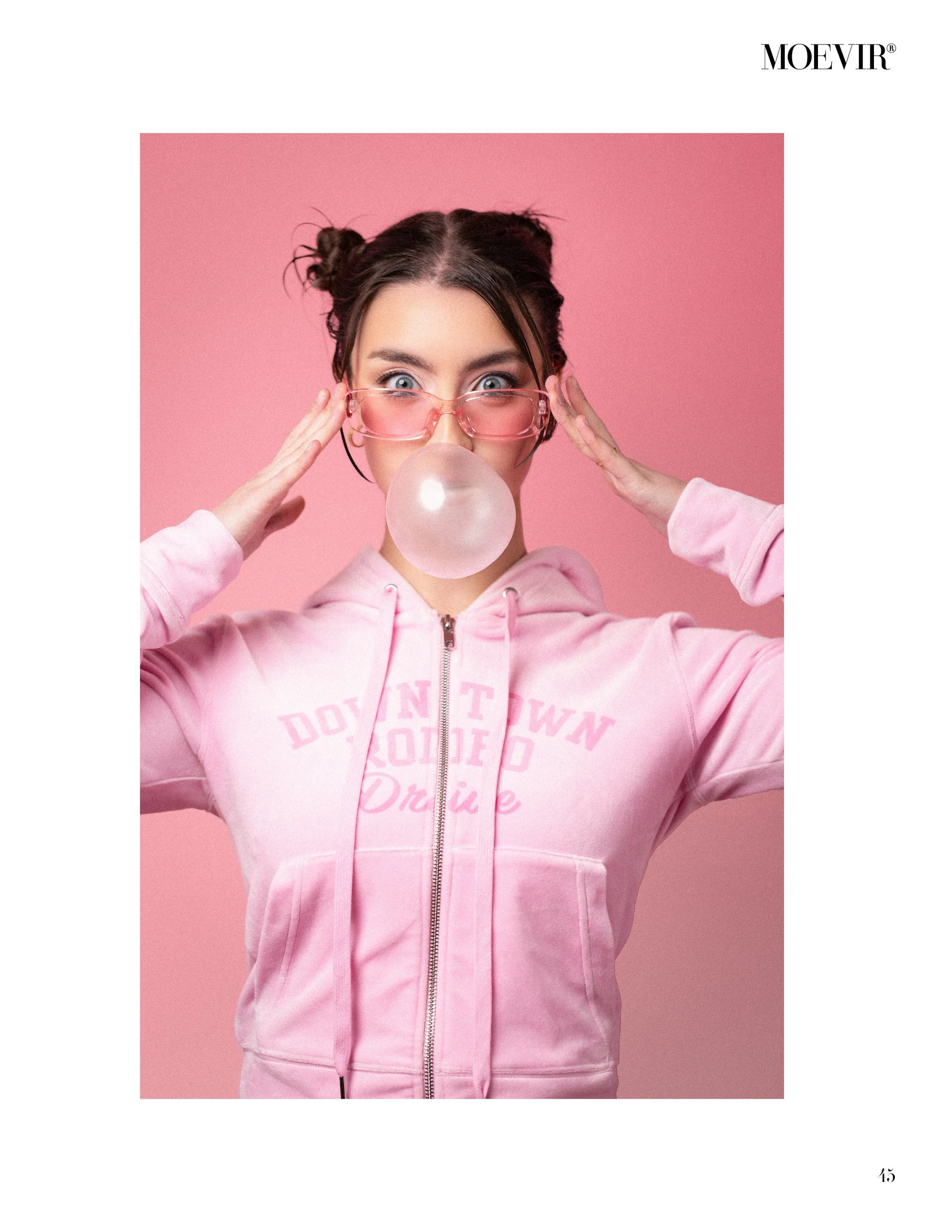 Young woman with dark hair styled in two buns, wearing pink glasses and a pink hoodie, blowing a bubble gum bubble against a pink background.
