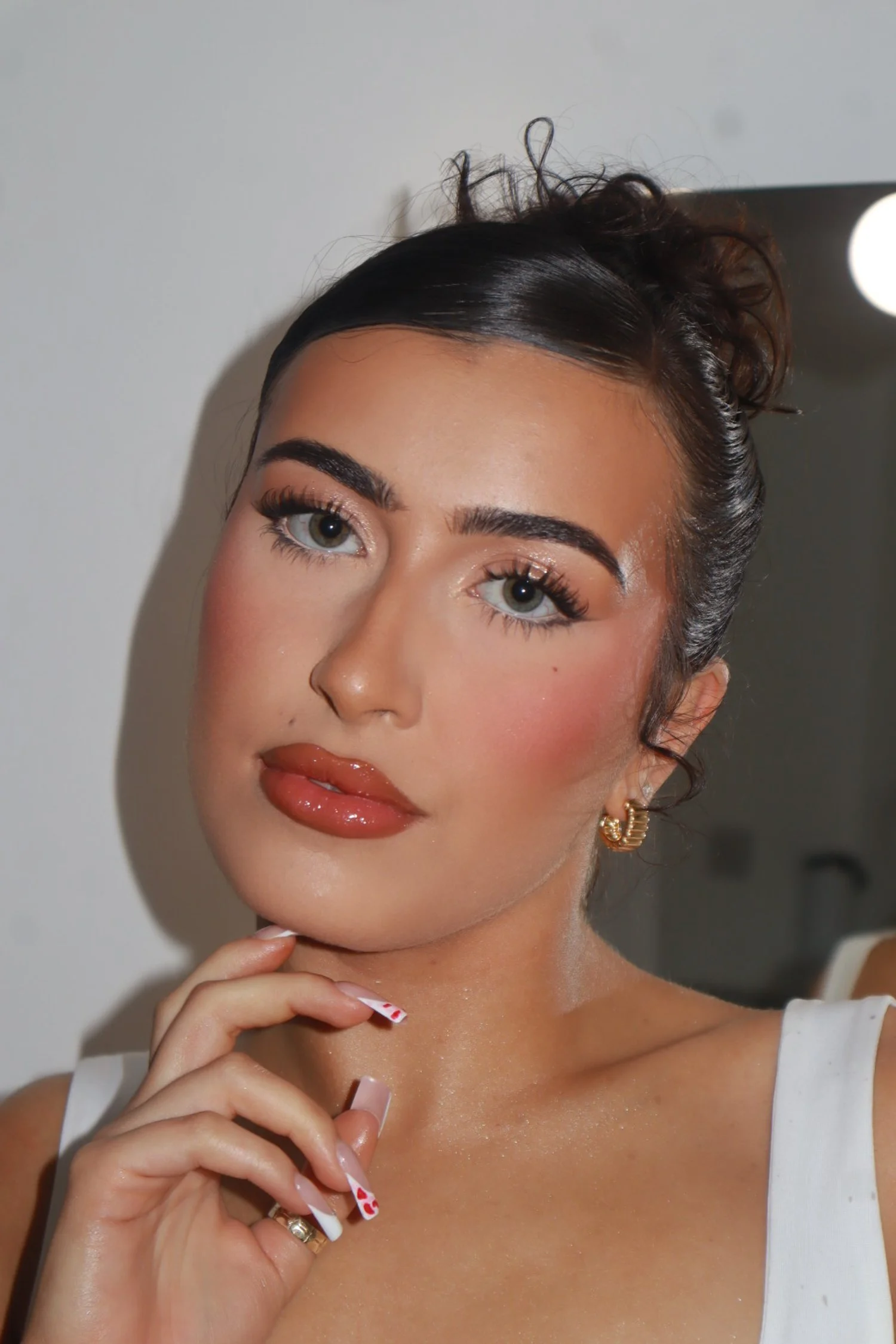 Close-up of a young woman with dark hair in a bun, green eyes, makeup, gold earrings, and a white top, posing with her hand near her chin.