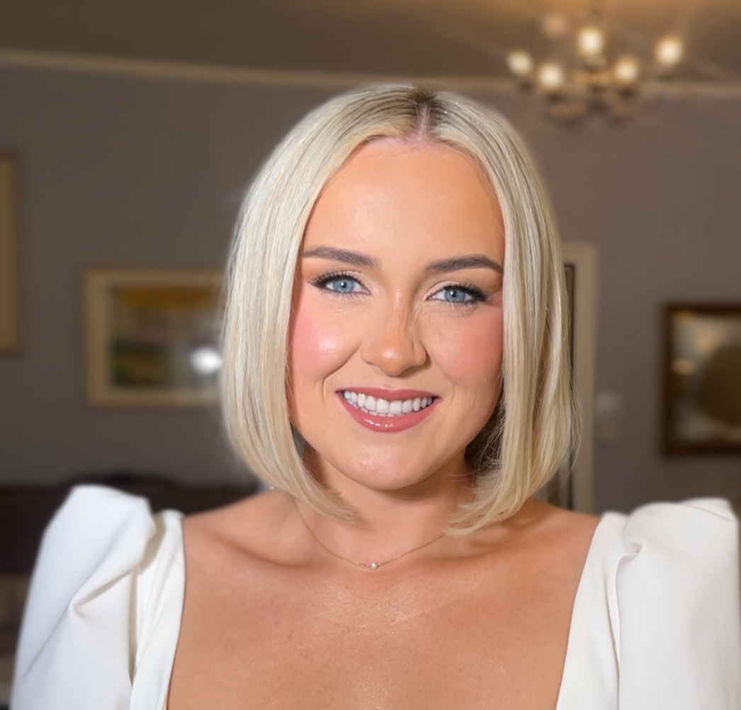 A woman with medium-length blonde hair, blue eyes, and a bright smile, wearing a white top or dress, standing in a well-lit living room with framed artwork on the walls and a chandelier on the ceiling.