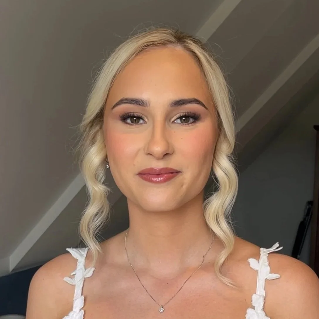 A young woman with blonde wavy hair and light makeup wearing a white top with flower details, posing indoors against a neutral background.