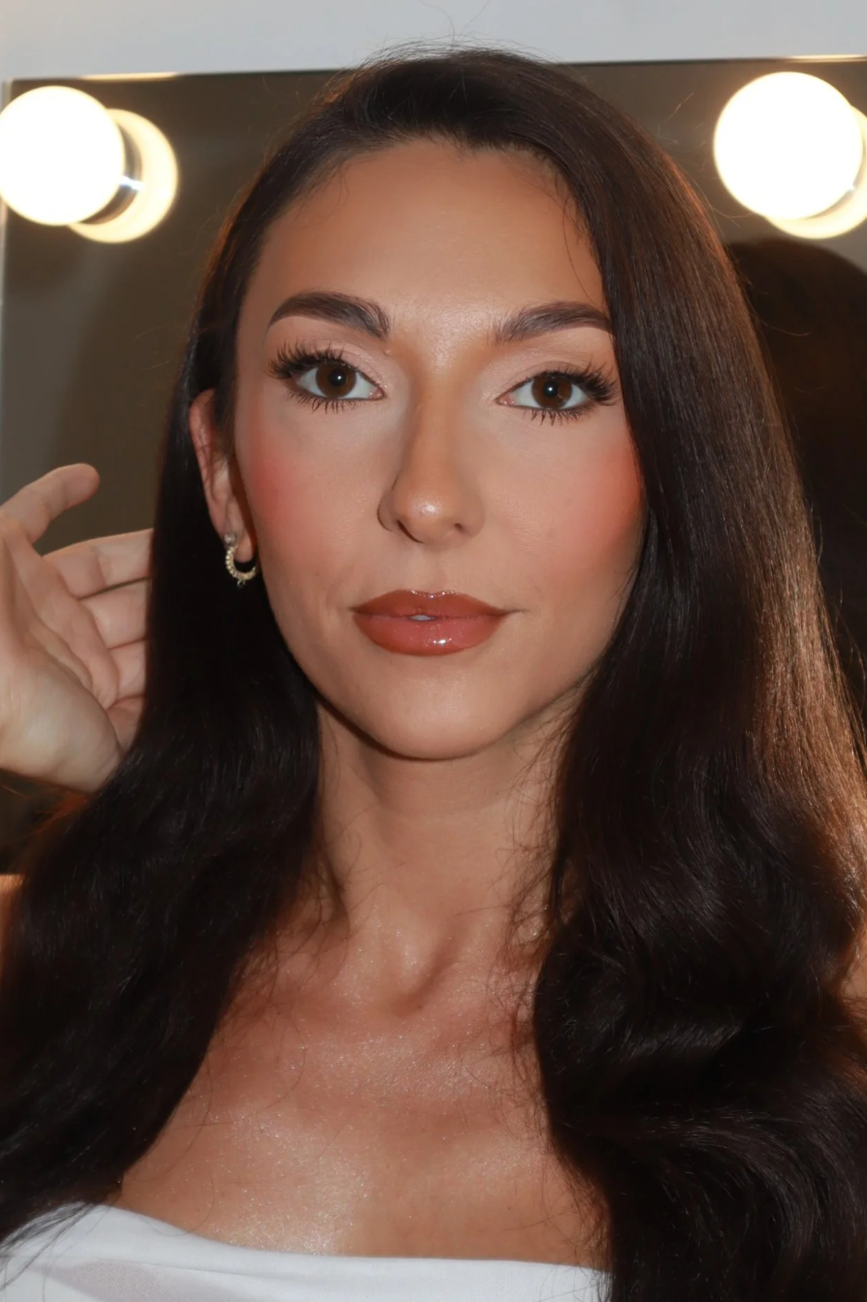 Close-up of a woman with long dark hair, soft glam makeup, and hoop earrings, in front of a mirror with bright lights.
