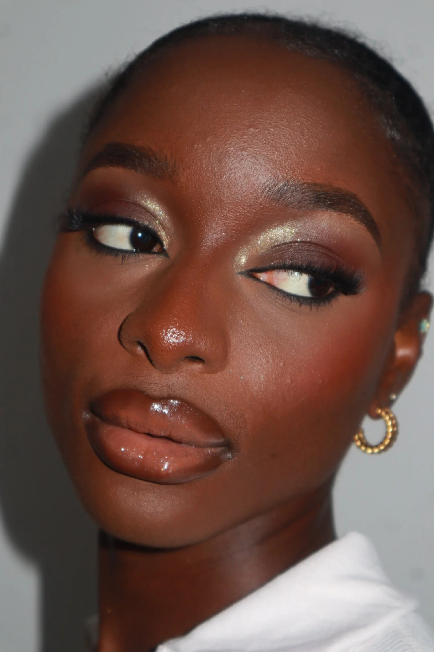 Close-up of a woman with dark skin and glossy lips, wearing occasion makeup, gold hoop earrings and makeup with shimmery eyeshadow.