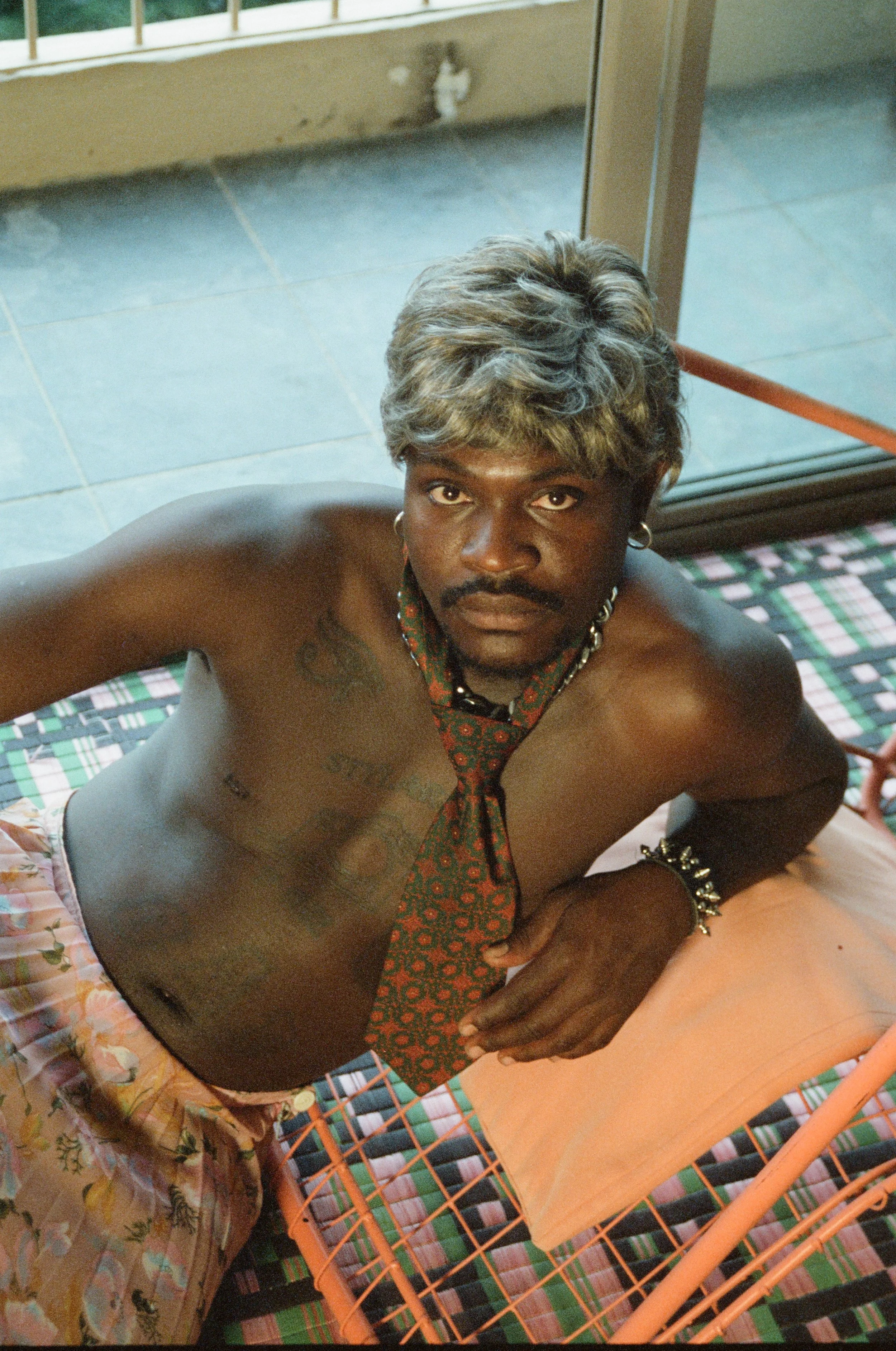 A shirtless man with tattoos and a colorful tie, lying on a bed, looking directly at the camera, with a serious expression.