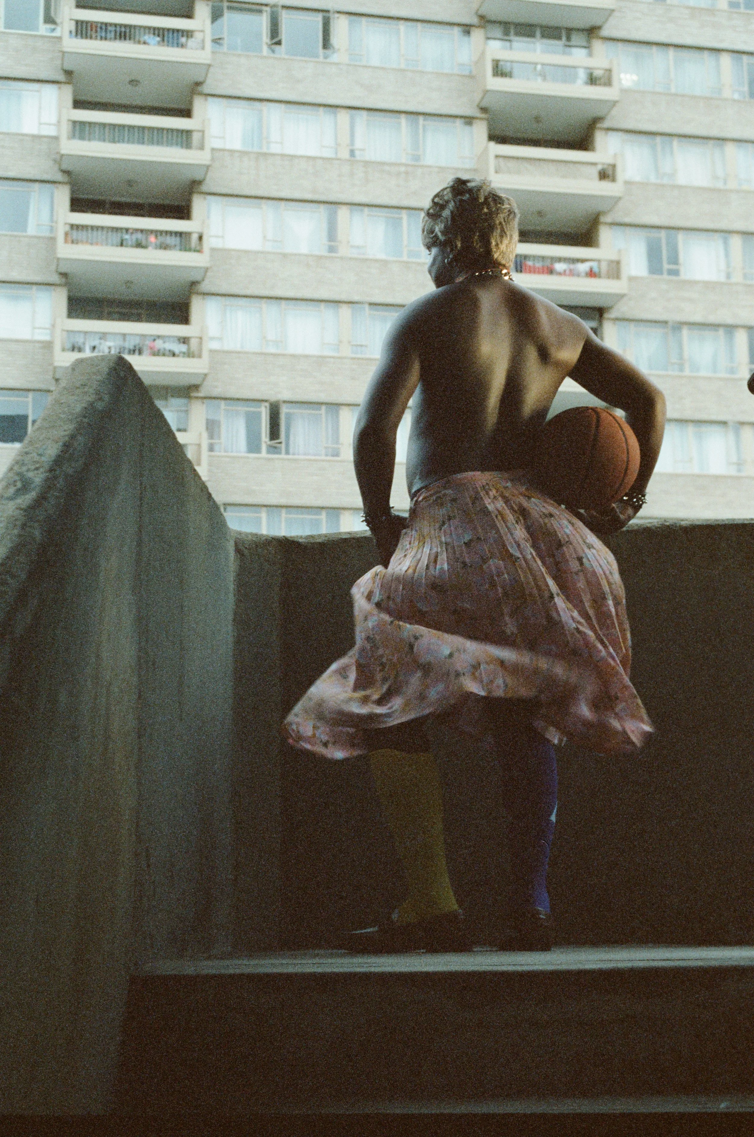 A shirtless man with curly hair, wearing a colorful skirt and rain boots, holds a basketball while standing on a balcony with a modern apartment building in the background.