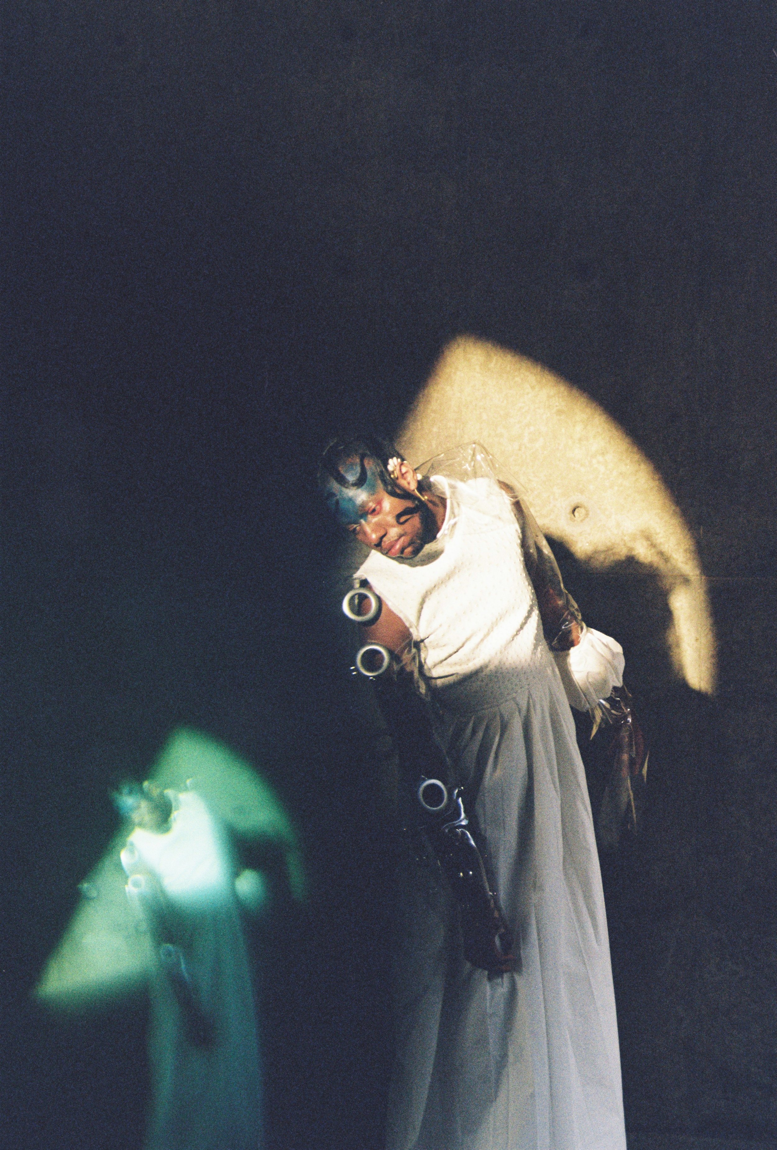 A woman taking a selfie in a mirror with a dark background and a spotlight on her face, wearing a white top and long skirt, with jewelry and tattoos visible.