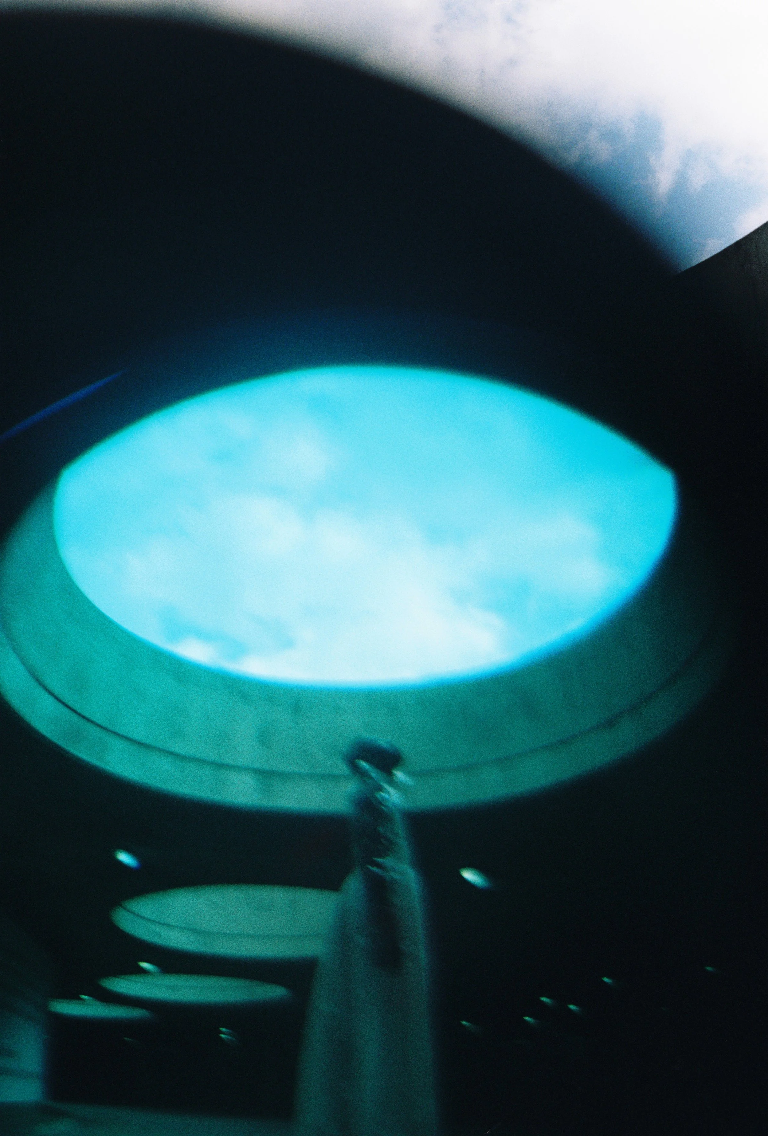 Interior view of a modern building with circular ceiling lights, with one prominently in the foreground showing a sky with clouds reflected on its surface.