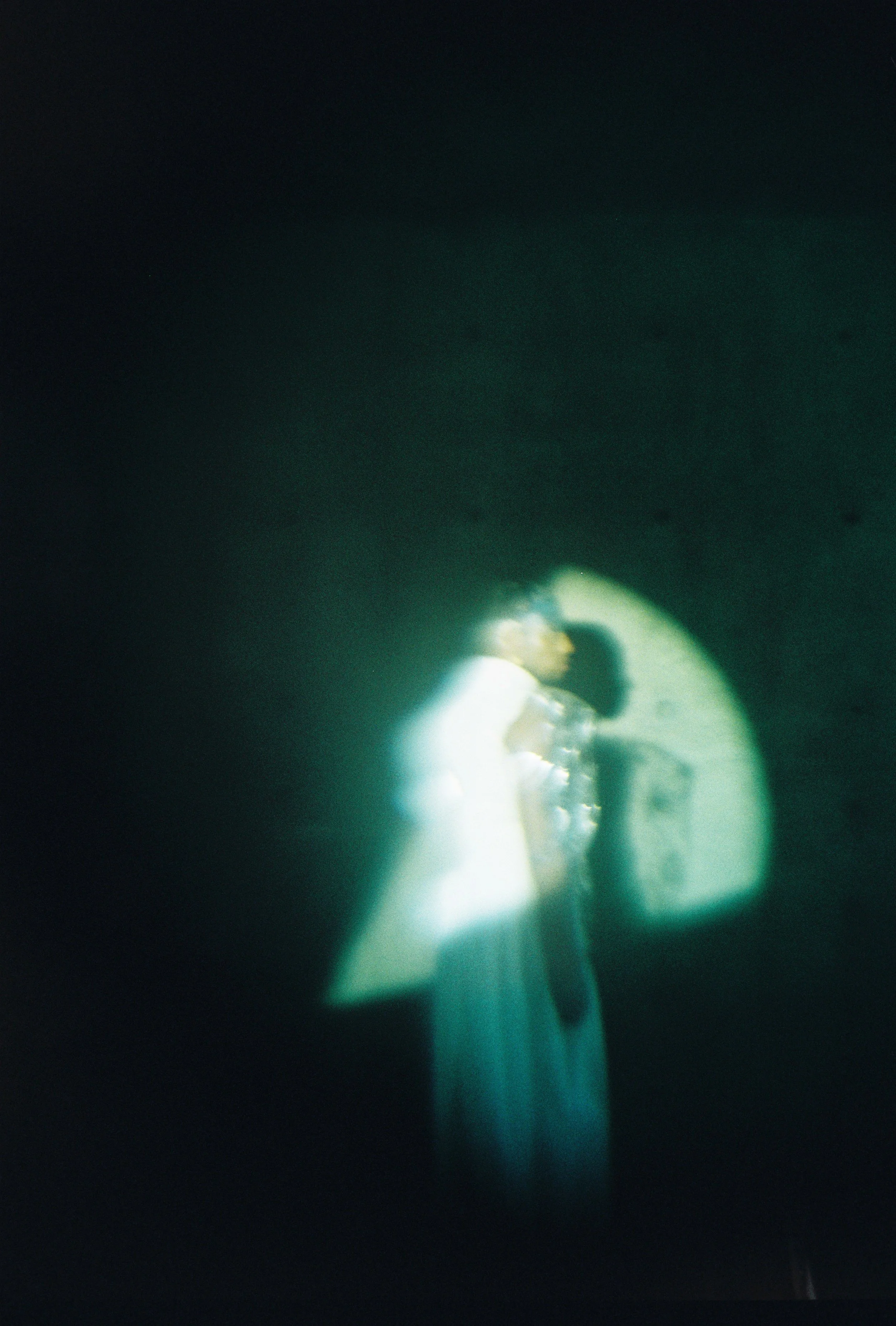 A blurry, glowing scene of a person in a white dress standing in front of a wall with a door or window, with indistinct objects or features around.