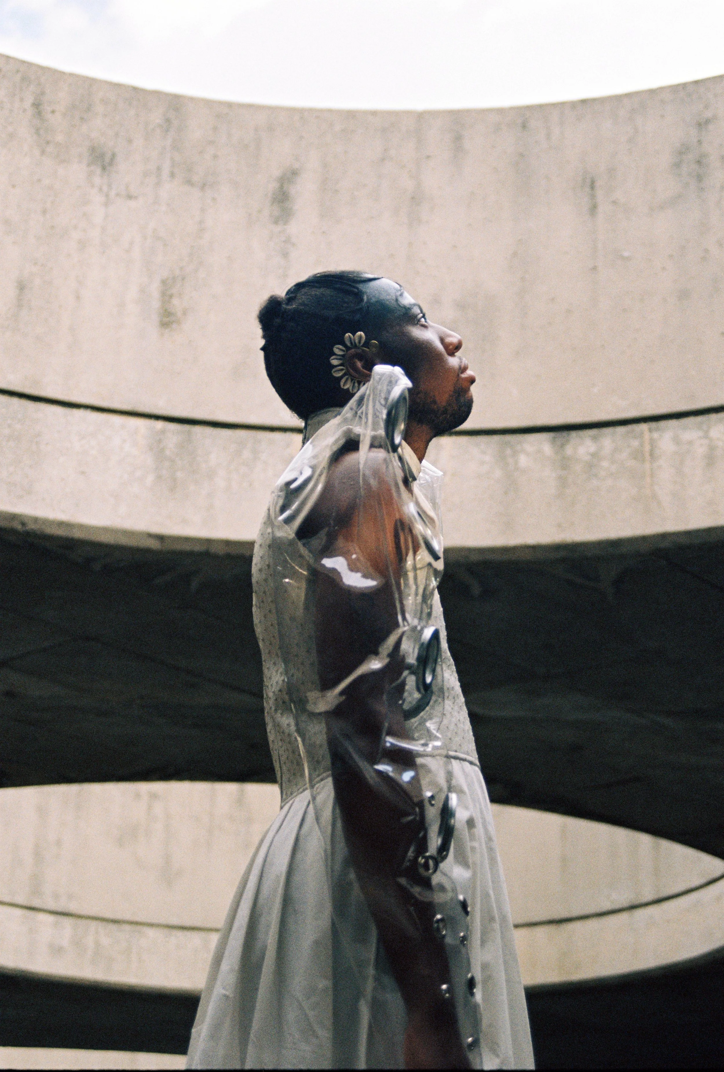 A person with styled hair and jewelry, wearing a transparent plastic outfit, standing against a concrete structure.