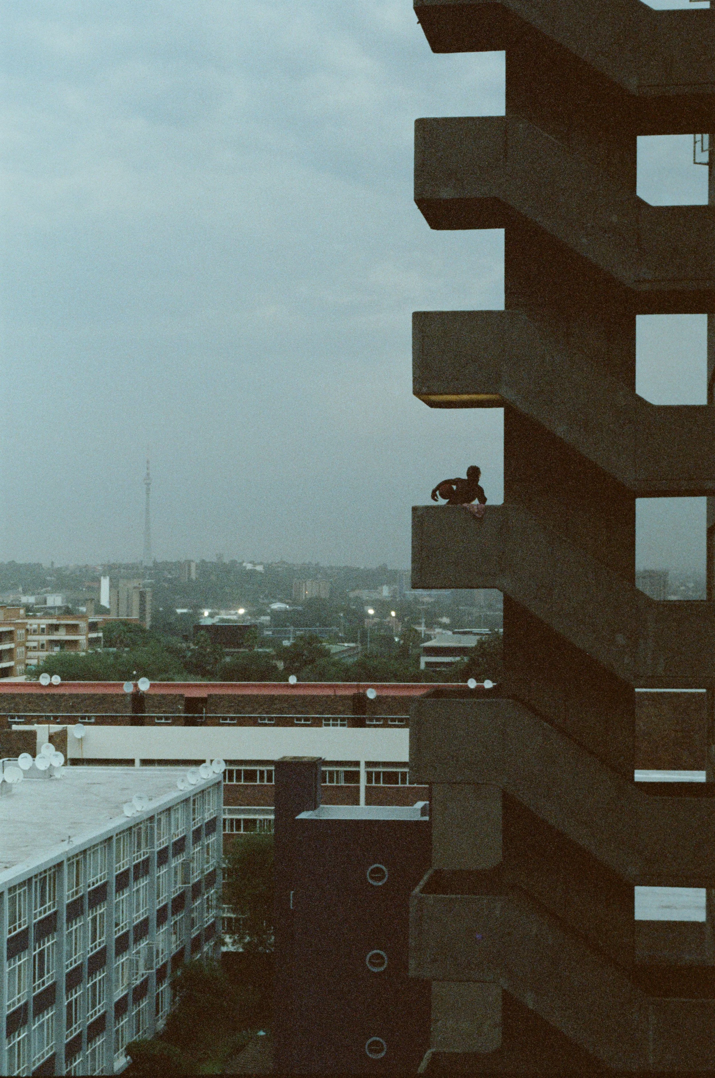 A silhouette of a person on the balcony of a tall apartment building, overlooking a cityscape on a cloudy day.