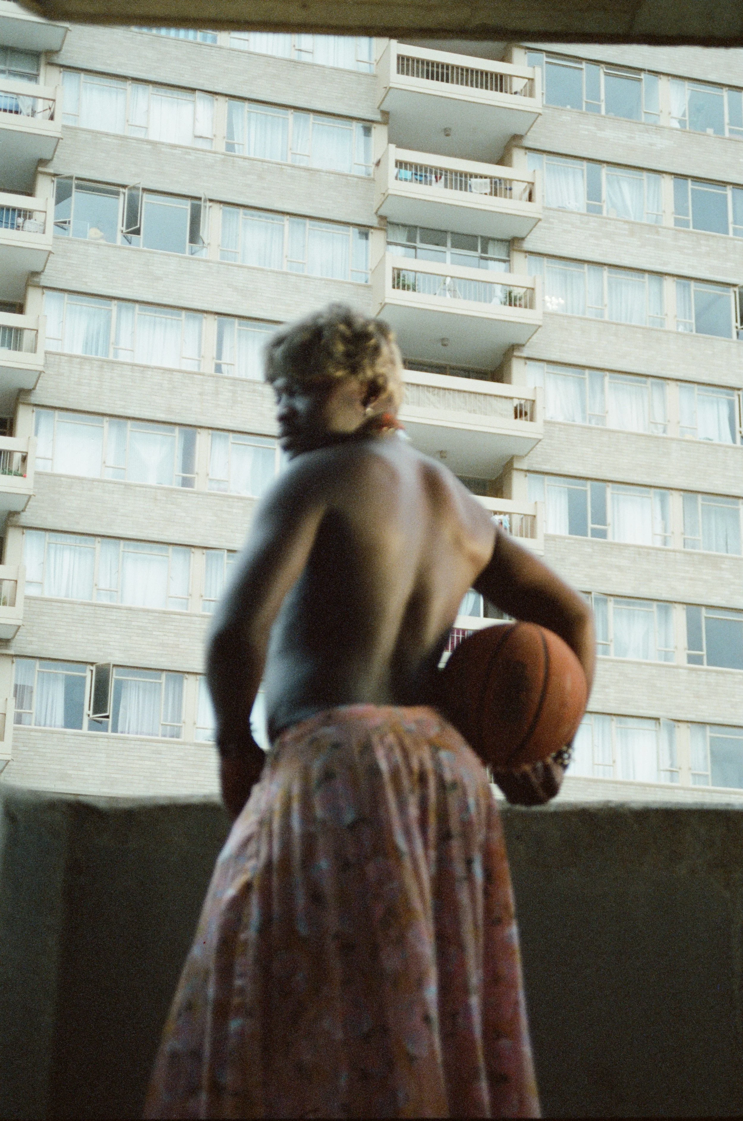 A shirtless young boy with curly hair holding a basketball on a balcony, with a large apartment building in the background.