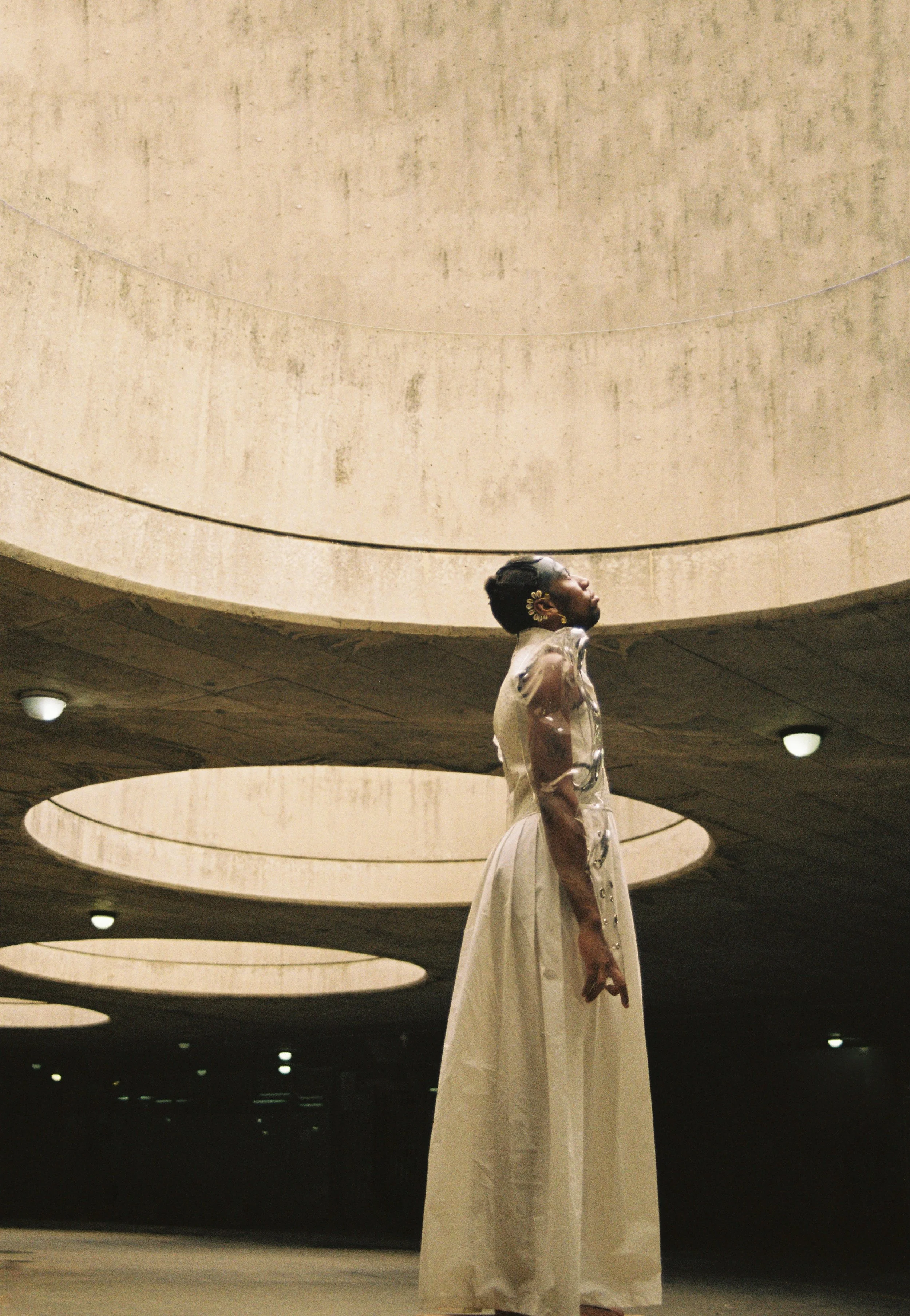A person standing under a concrete parking garage ceiling with circular cutouts, wearing a clear plastic shirt and long white skirt, with one hand hanging down and the head inclined upward.