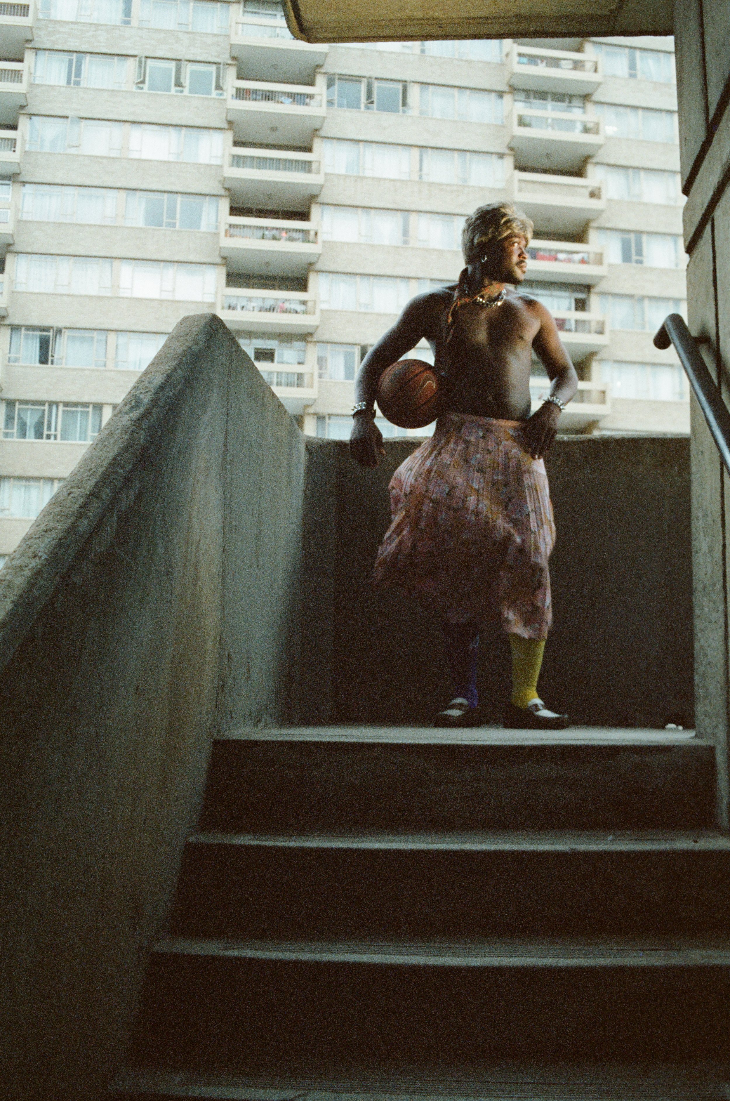 A shirtless man holding a basketball standing on stairs of an outdoor concrete balcony in front of a tall apartment building.