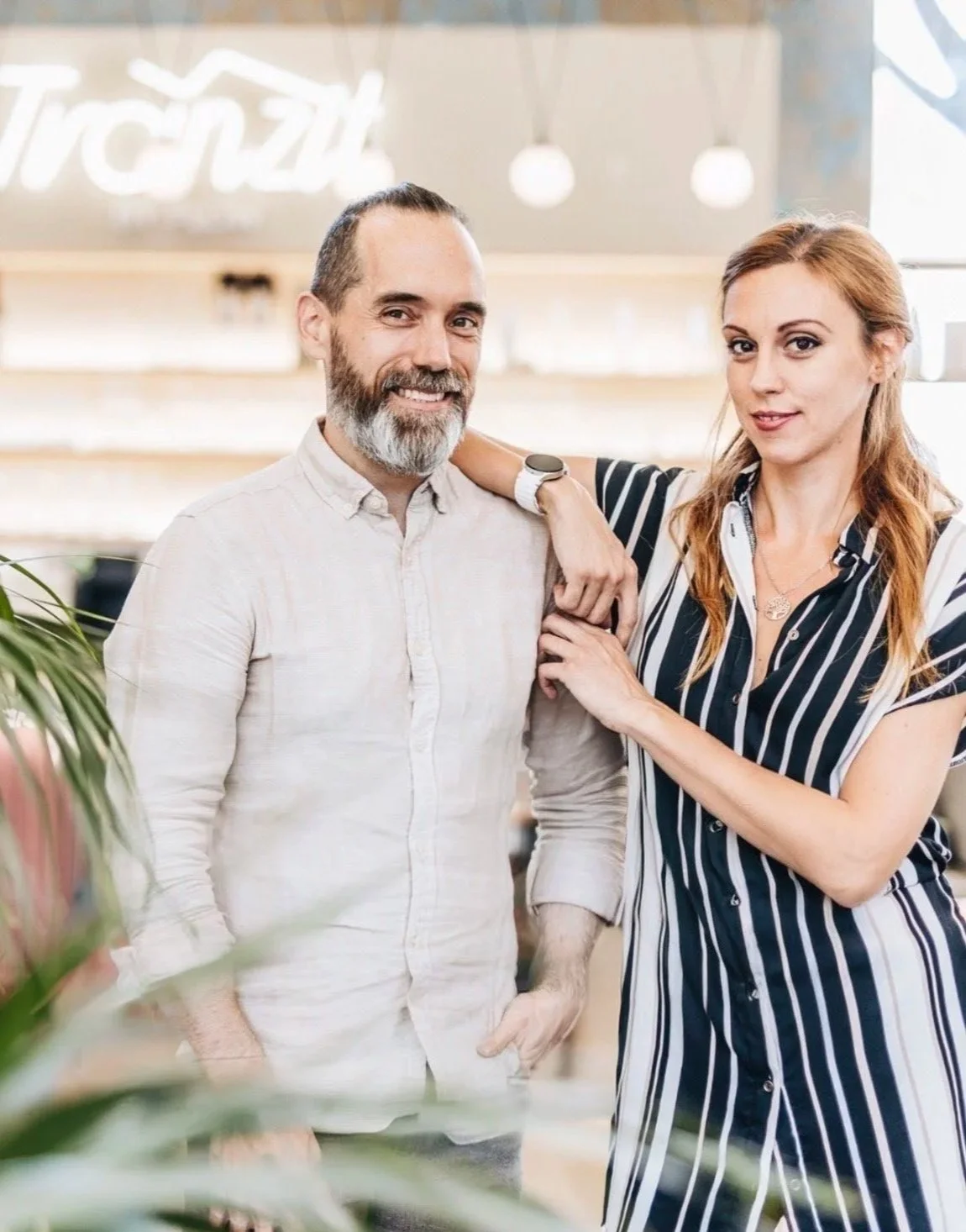 A man and woman standing close together inside a well-lit modern office or cafe, smiling at the camera. The man has a beard and gray hair, wearing a light-colored button-up shirt. The woman has long reddish hair, wearing a black and white striped shirt, with her arm draped over the man's shoulder.
