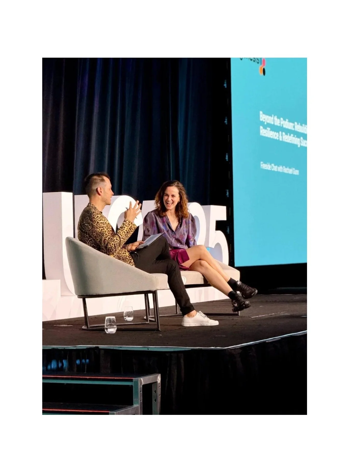 Raygun (Dr Rachael Gunn), wearing a purple patterned blouse and pink skirt, sitting on stage in an armchair next to a male interviewer in a leopard-print top, mid-conversation during the ALUCA 20th Anniversary Conference, October 2025.