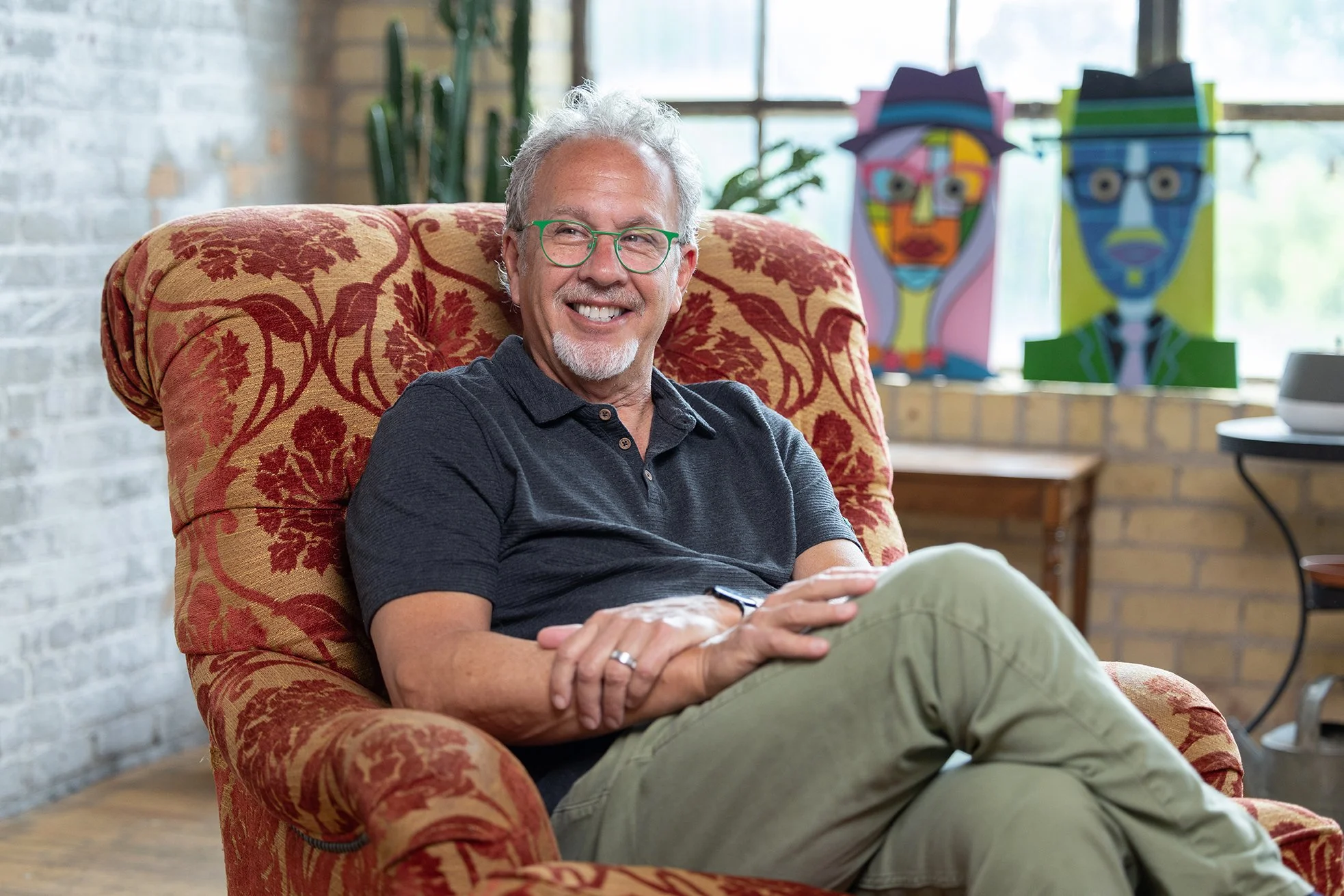 Smiling man with gray hair and green glasses sitting in a patterned armchair, with colorful abstract art in the background.